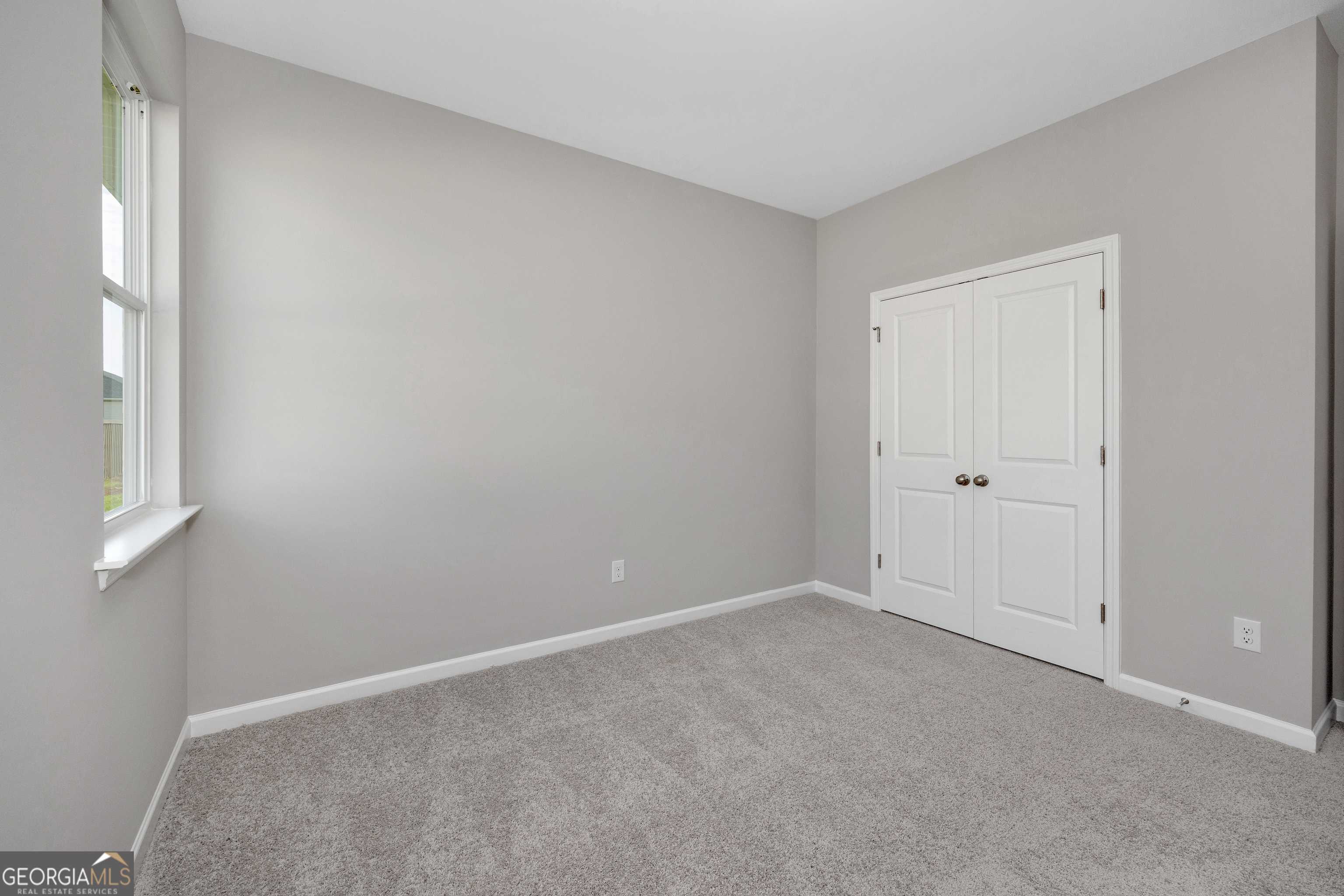 Empty bedroom with gray walls, plush carpet, double white closet doors, and large window in Evermore Homes The Luna, Ivy Glen, Perry, Georgia