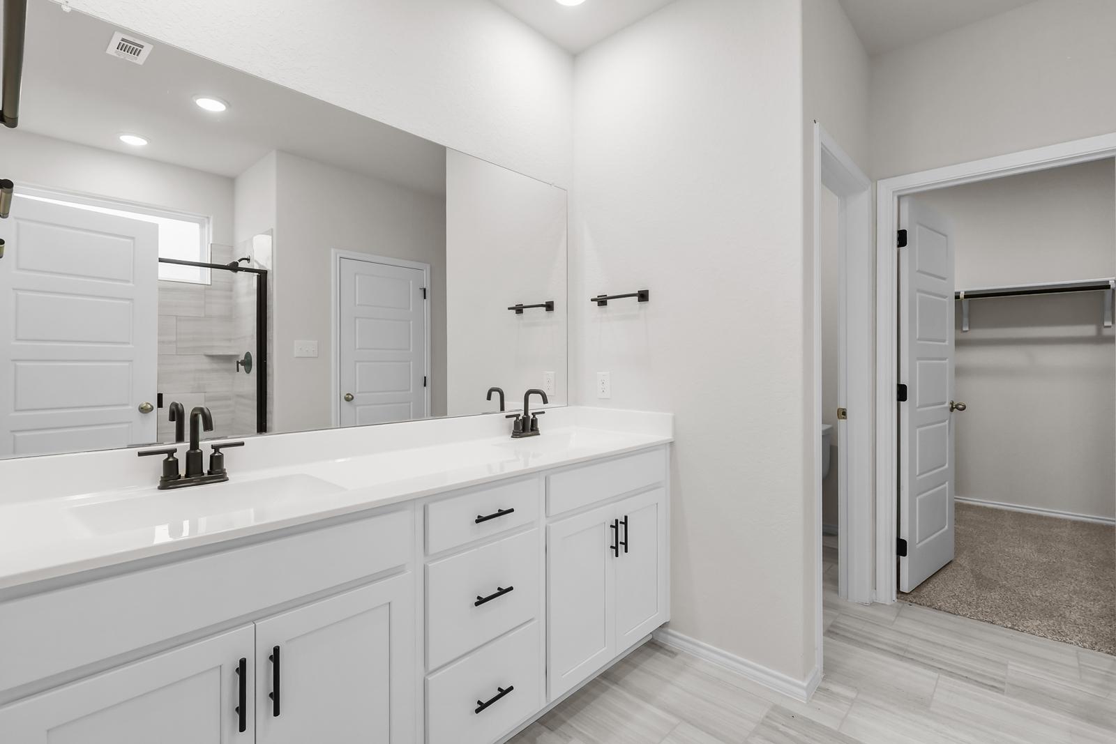 Modern primary bathroom in The Sequoia C featuring white dual-sink vanity, matte black fixtures, and walk-in closet.