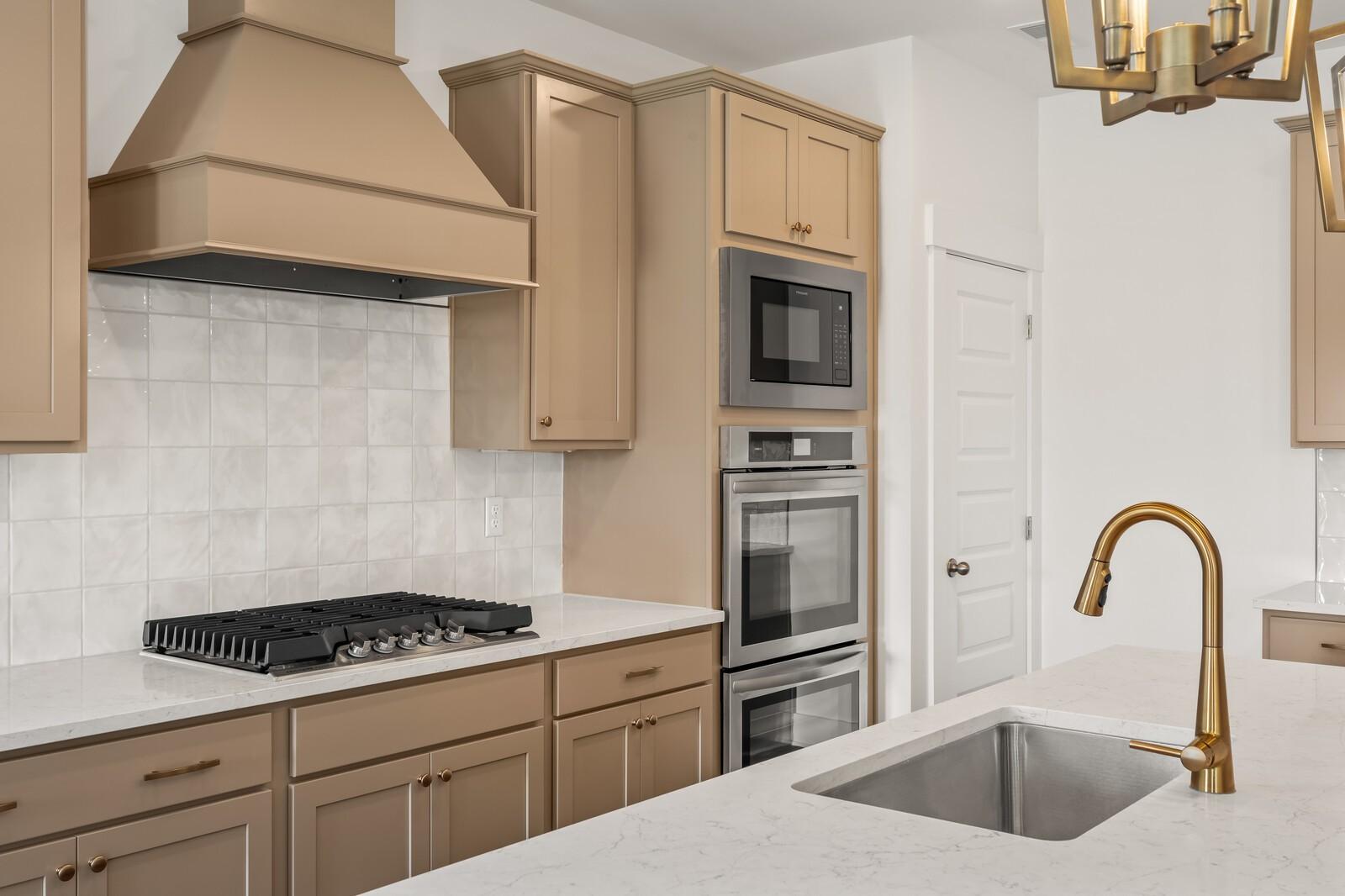 Modern kitchen featuring beige shaker cabinets, white subway tile backsplash, gas cooktop, double ovens, and gold faucet in Davidson Homes The Ash A, Gallatin, TN