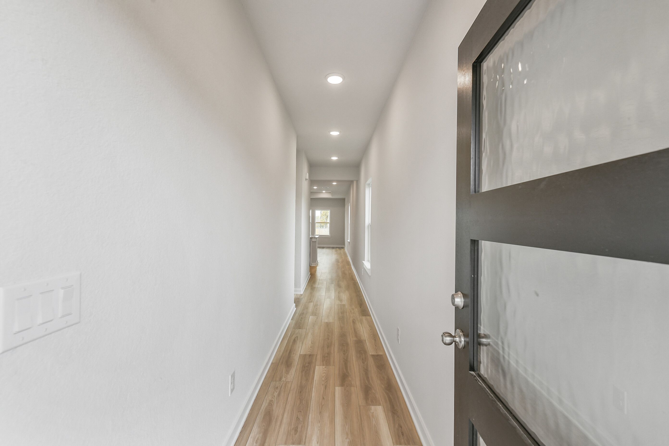 Long hallway with light oak floors, white walls, recessed lights, and frosted glass door in The Colorado G home, Magnolia Texas