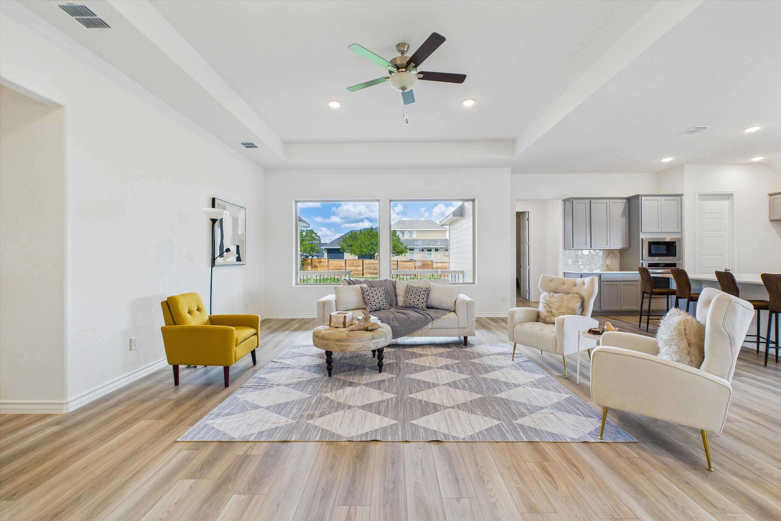 Bright open living room with ceiling fans, plush sofa, armchairs, and hardwood floors open to white kitchen in Davidson Homes The Summerlin C, Castroville, Texas