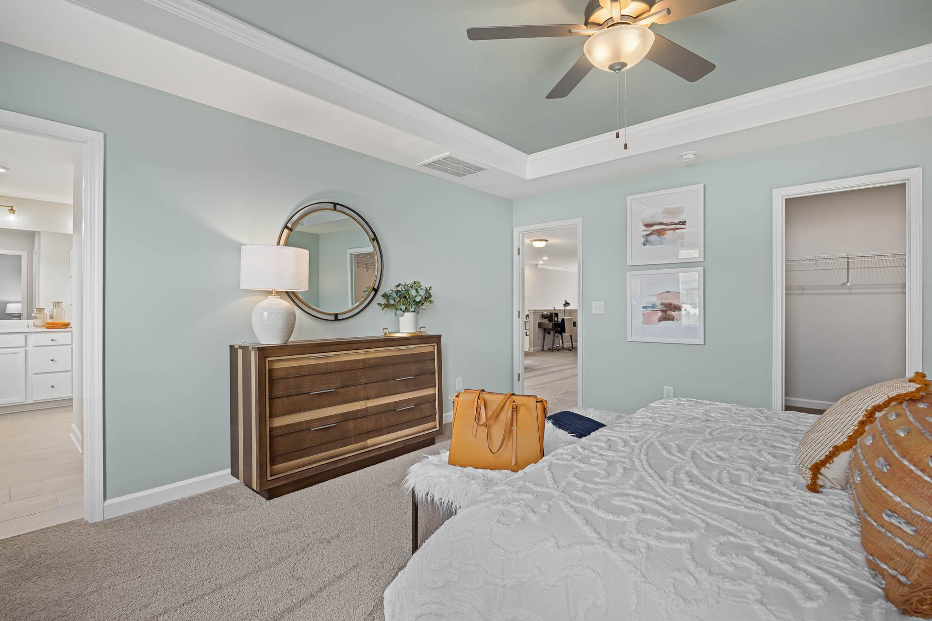 Spacious primary bedroom at Beverly Place in Four Oaks NC with light blue walls, wooden dresser, mirrored vanity, and plush white king bed