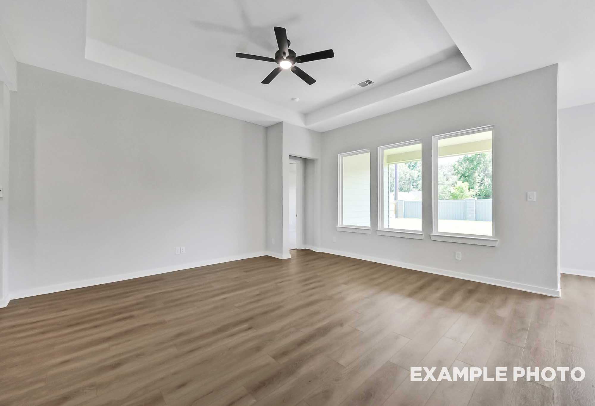 Spacious open living room in The Sequoia home with tray ceiling fan, light gray walls, and large triple windows overlooking greenery