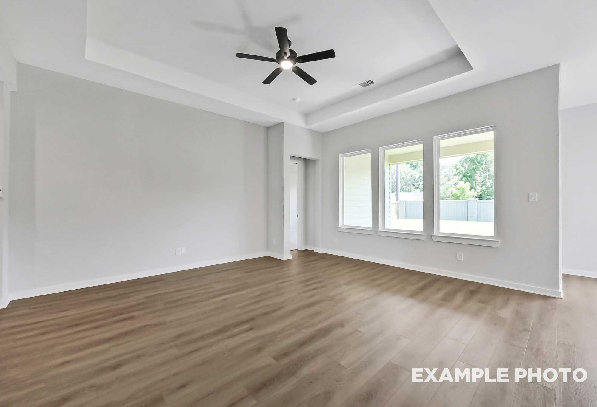 Spacious open living room in The Sequoia home with tray ceiling fan, light gray walls, and large triple windows overlooking greenery