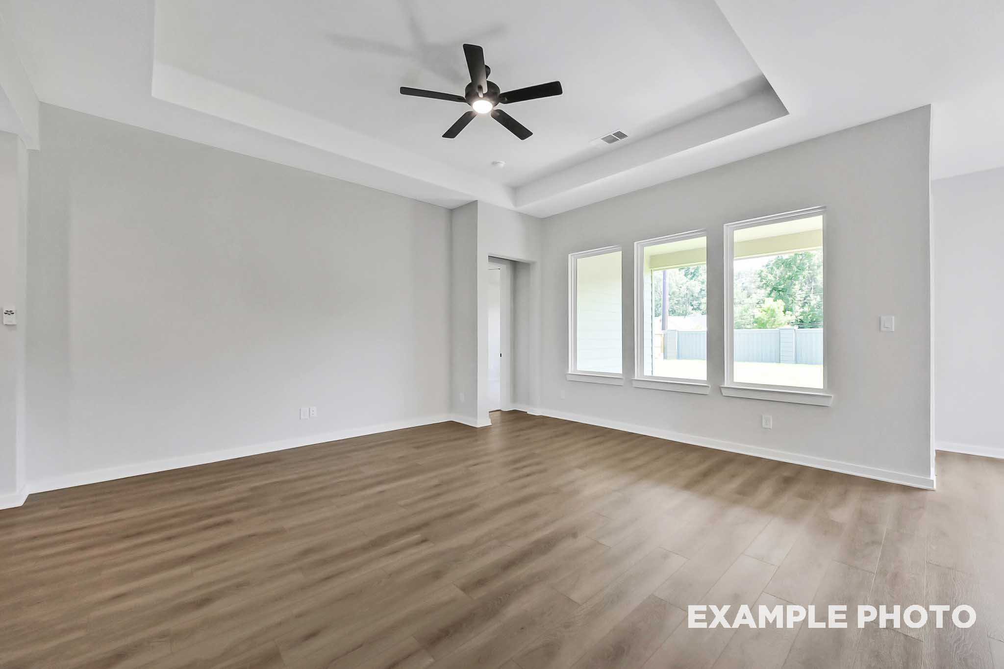 Spacious open living room in The Sequoia home with tray ceiling fan, light gray walls, and large triple windows overlooking greenery
