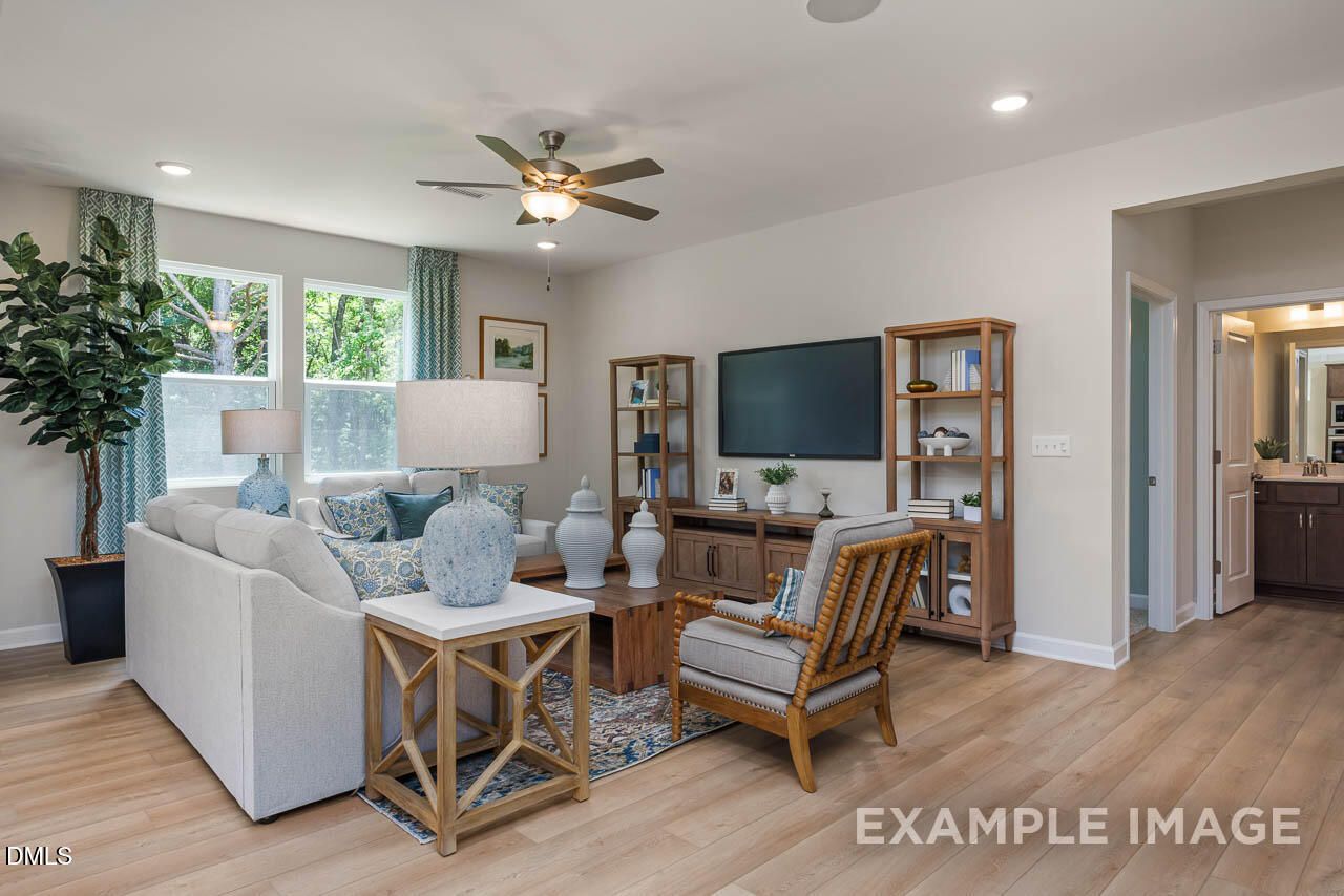 Cozy living room with white sofa, wooden coffee table, rattan chair, TV stand, potted plants, and large windows in Davidson Homes The Willow B, Lillington, NC