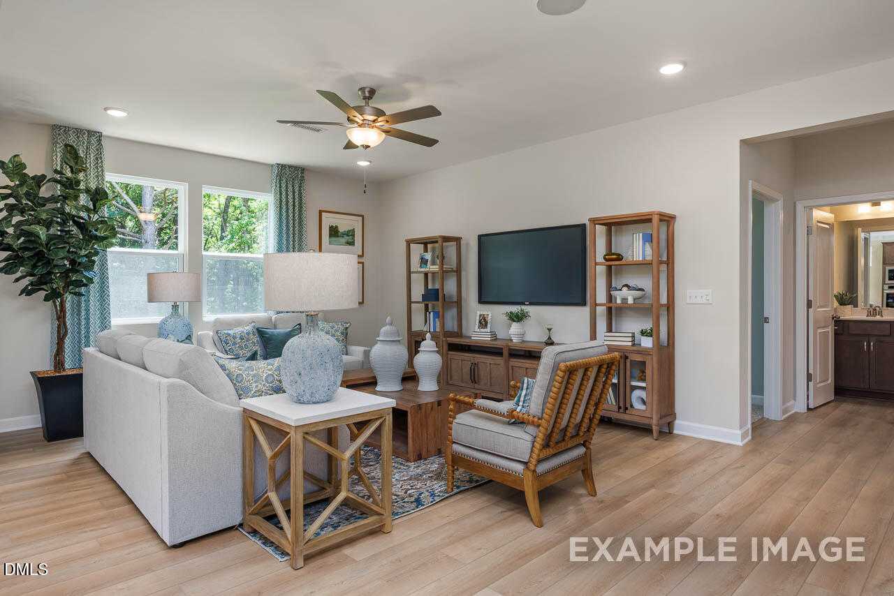 Cozy living room with white sofa, wooden coffee table, rattan chair, TV stand, potted plants, and large windows in Davidson Homes The Willow B, Lillington, NC