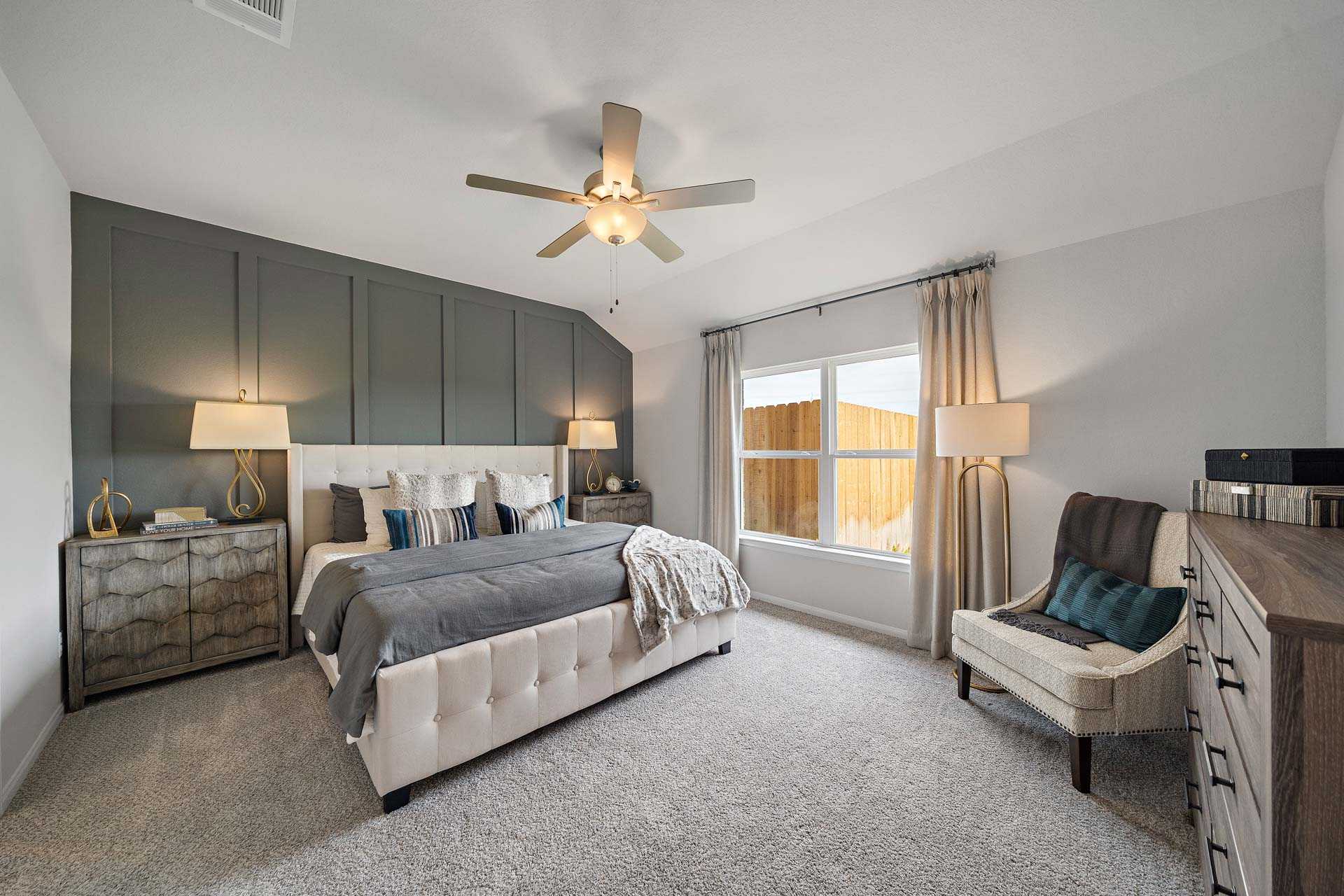 Spacious master bedroom in Sunterra Katy TX with gray accent wall, tufted king bed, ceiling fan, and window by Davidson Homes