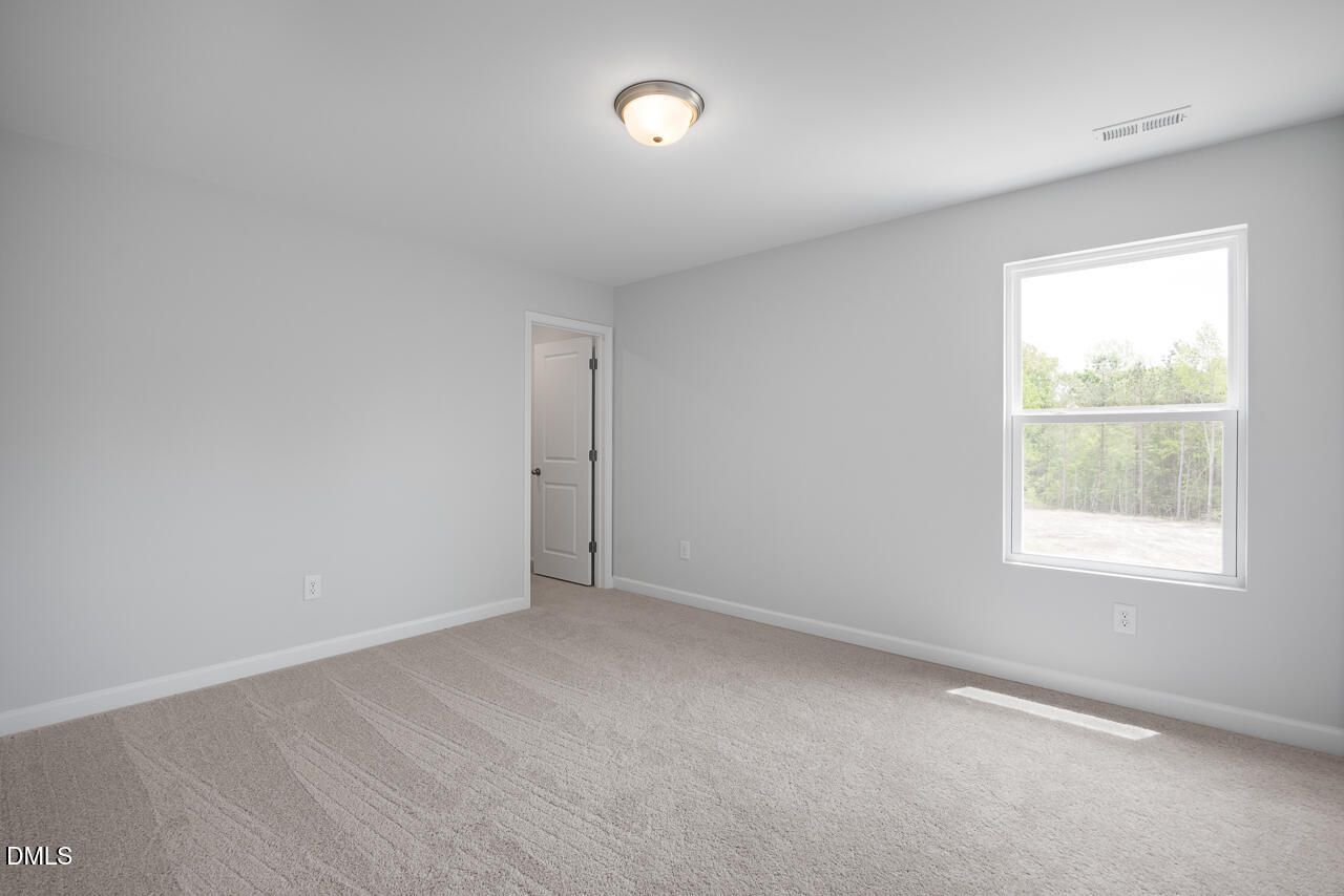 Spacious empty bedroom with light gray walls, large window overlooking trees, carpeted floor in Davidson Homes The Hickory II B, Zebulon NC