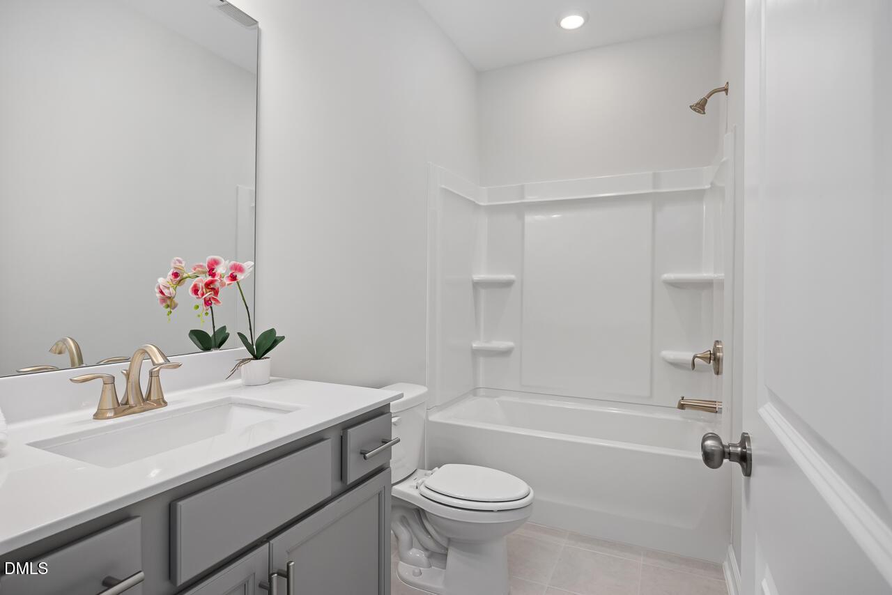 Bright guest bathroom with white tub-shower, quartz vanity, gold fixtures, and orchids in Davidson Homes The Daphne D, Lillington, NC