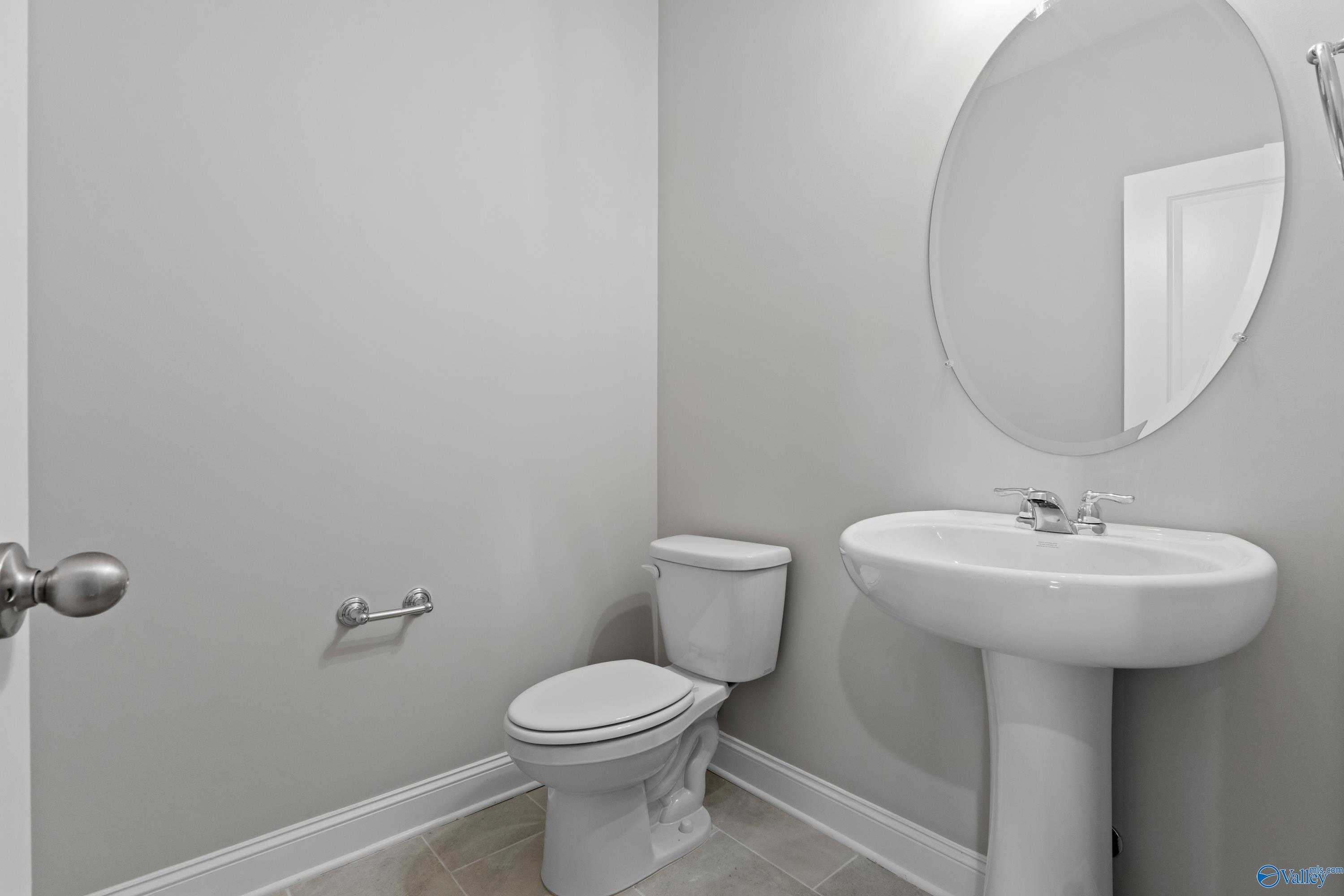 Elegant powder room featuring round mirror, pedestal sink, and toilet in soft gray tones in Davidson Homes The Finleigh, Meridianville, Alabama