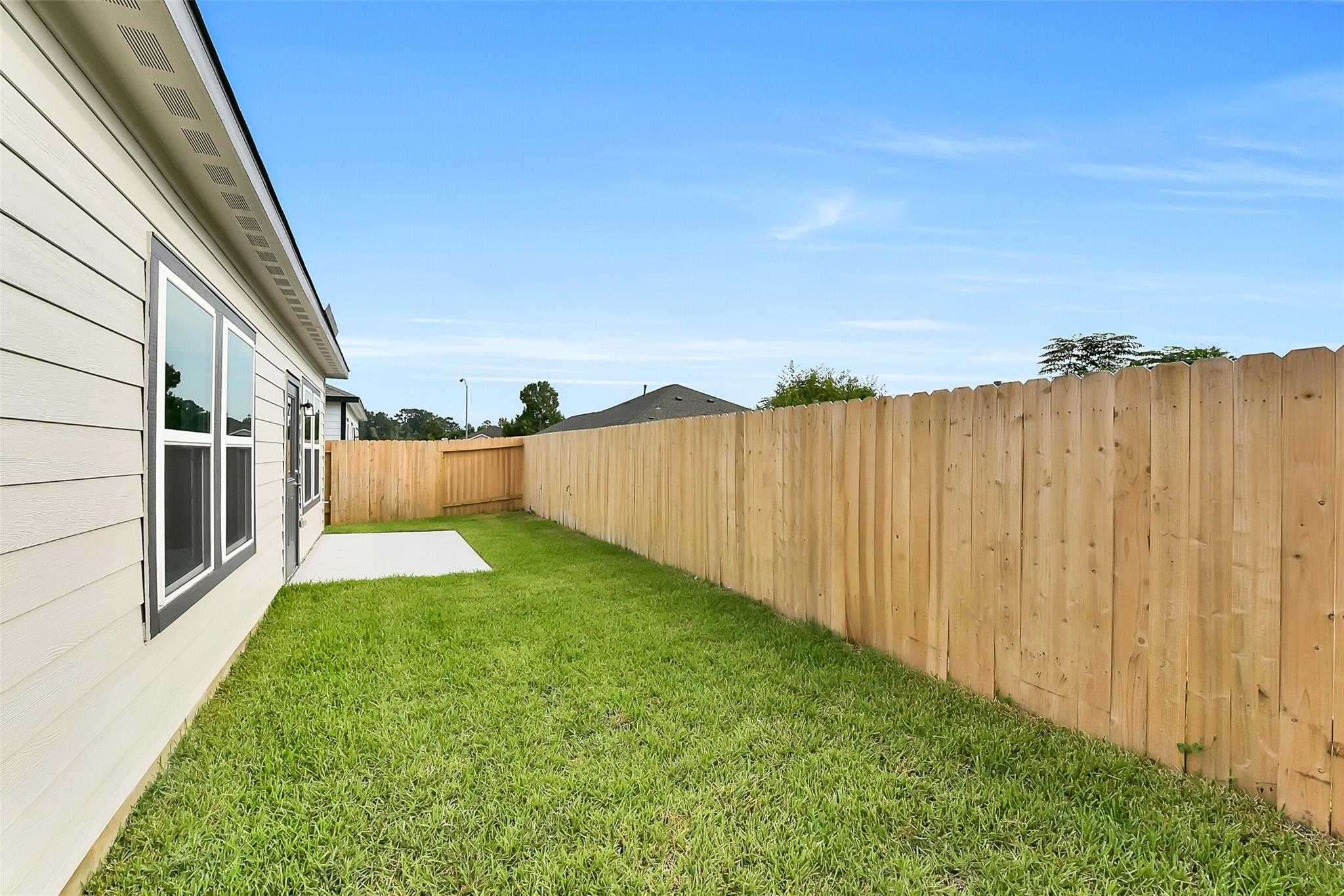 Private backyard with wooden privacy fence and lush green lawn beside beige single-story The Frio G home in Dayton, Texas