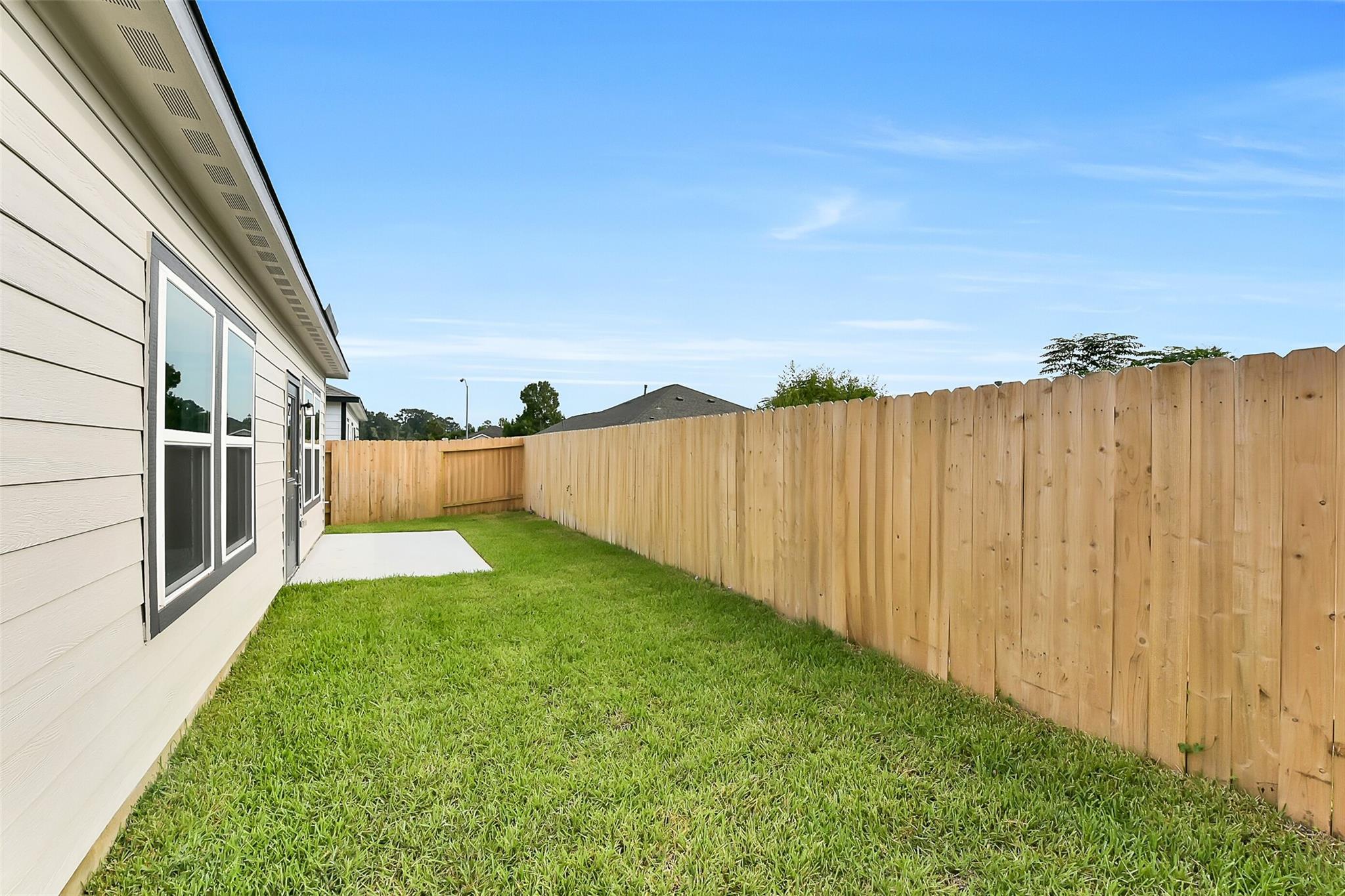 Single-story beige home side view with wooden privacy fence, lush green lawn, and concrete slab in The Villages at WestPointe, Dayton, Texas