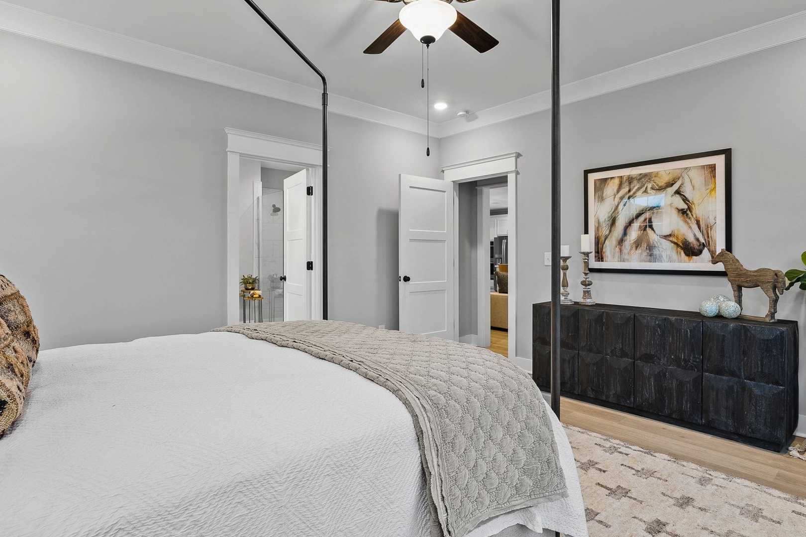 Spacious master bedroom in The Oxford by Evermore Homes featuring king canopy bed, ceiling fan, abstract horse art, and en-suite bath