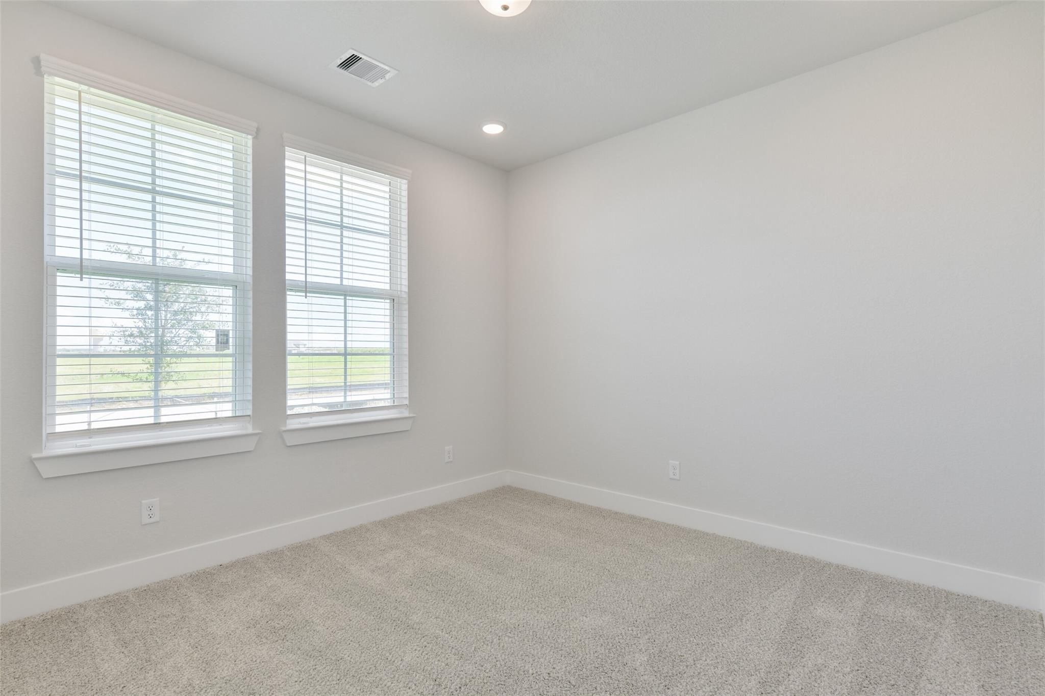 Bright empty bedroom with neutral gray walls, beige carpet, and large windows with blinds overlooking field in Davidson Homes The Edward A, Lago Mar, Texas City