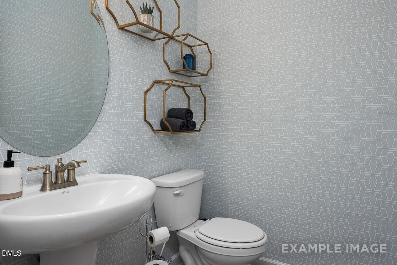 Modern powder room with blue geometric wallpaper, gold shelves with towels and plants, round mirror, pedestal sink, toilet in Davidson Homes The Magnolia B, Angier, NC