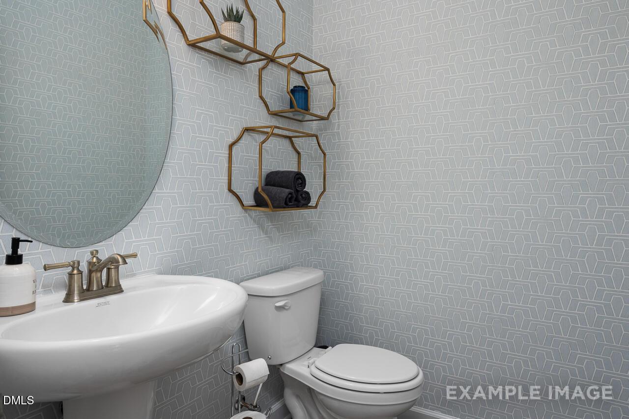 Modern powder room with blue geometric wallpaper, gold shelves with towels and plants, round mirror, pedestal sink, toilet in Davidson Homes The Magnolia B, Angier, NC