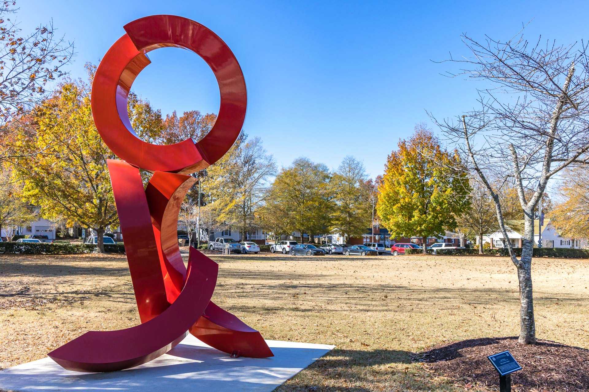 Abstract red metal sculpture in Sierra Heights park, Clayton NC amid fall foliage, grassy field, and distant homes