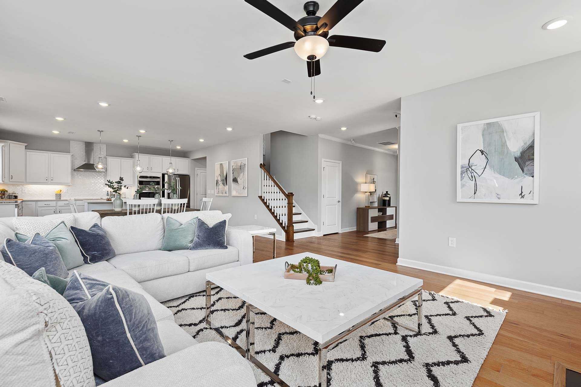 Open-concept living room at Highland Forest in Fuquay-Varina NC with white sectional sofa, marble coffee table, hardwood floors, and staircase