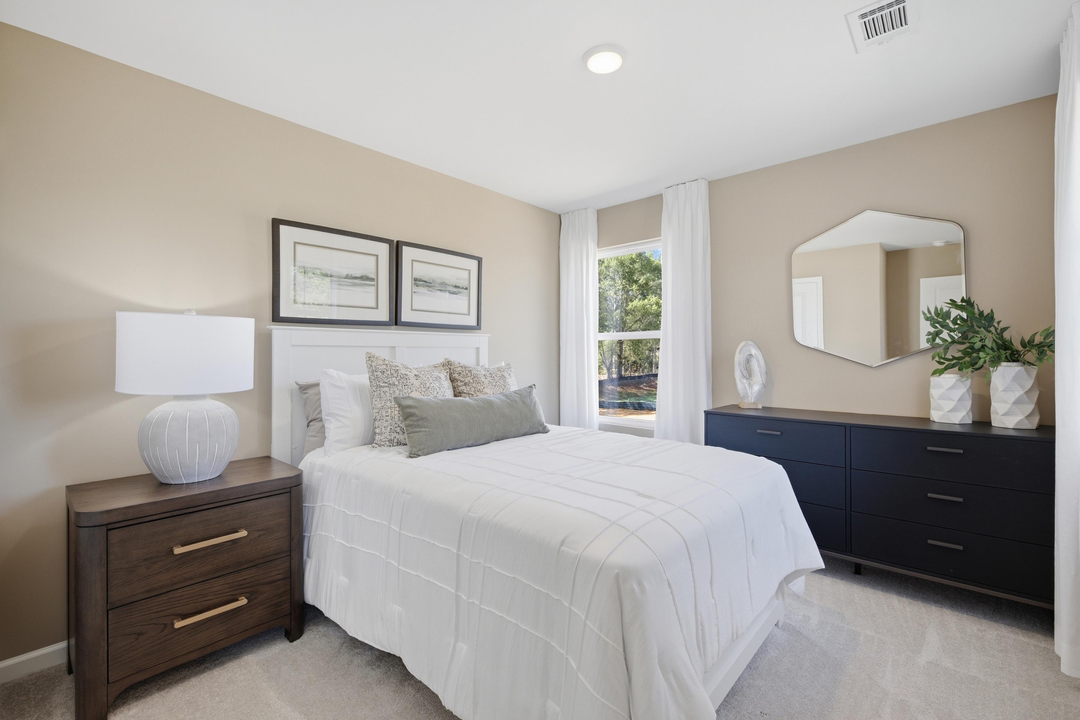Bright neutral bedroom with white bedding, wood nightstand, and navy dresser at Greene Farm by Davidson Homes in Cartersville, GA
