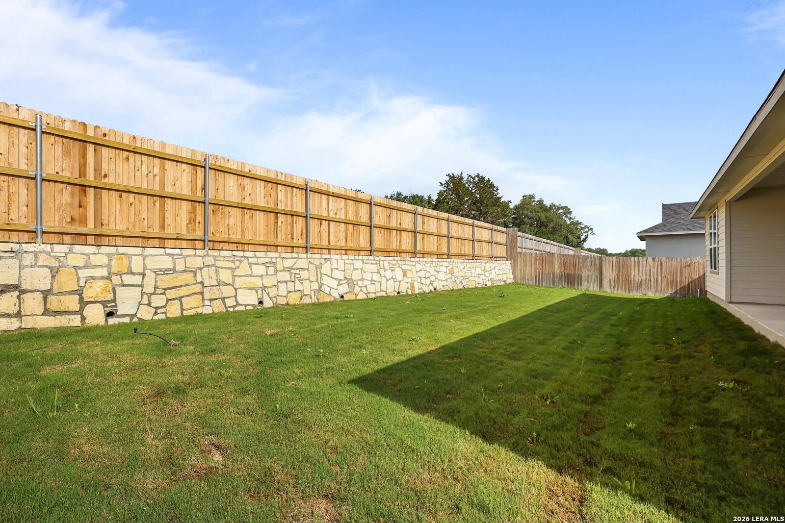 Private fenced backyard with lush green lawn and stone wall base in Davidson Homes The Lanier G, Castroville, Texas