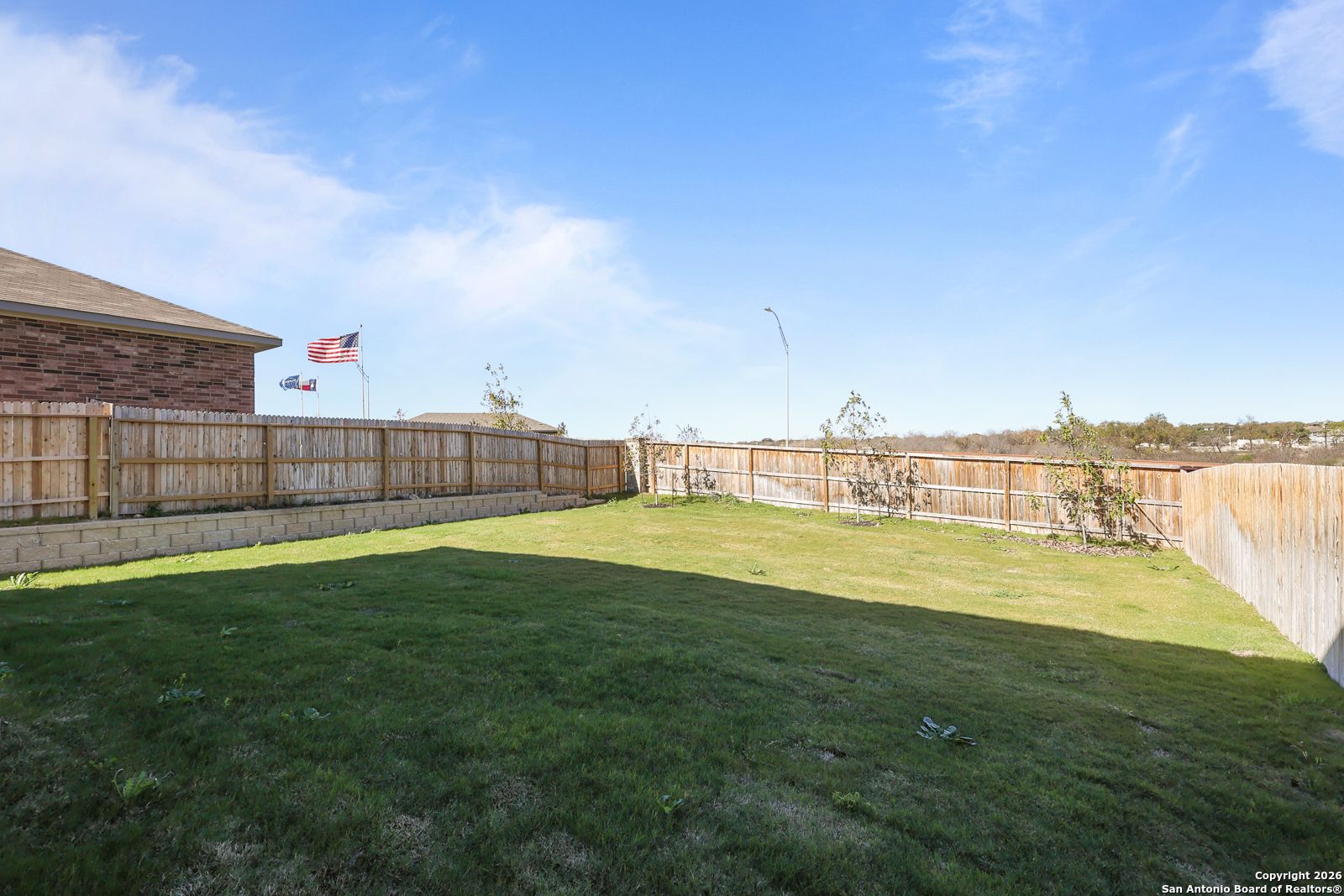 Private fenced backyard with lush green lawn, brick home exterior, and American flag in Davidson Homes The Murray K, Comanche Ridge, San Antonio, Texas
