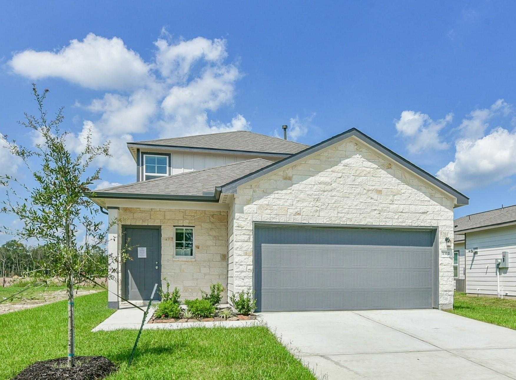 Two-story beige stone home with gray 2-car garage, front yard landscaping, and blue sky in The Villages at WestPointe, Dayton, Texas - Davidson Homes The Sabine E