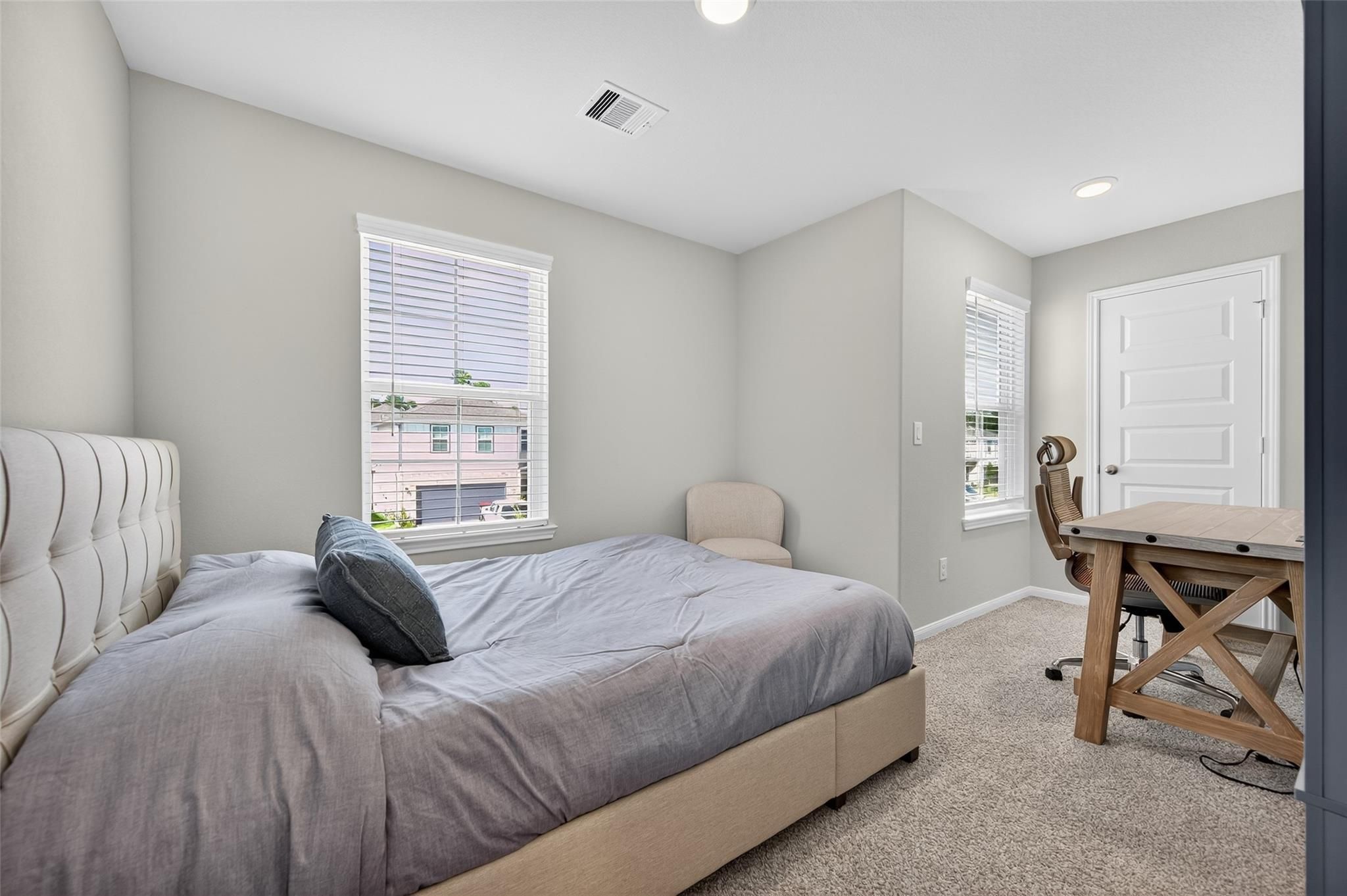 Inviting gray bedroom with tufted bed, wooden desk, armchair, and window views in Davidson Homes The Brazos E, Magnolia TX