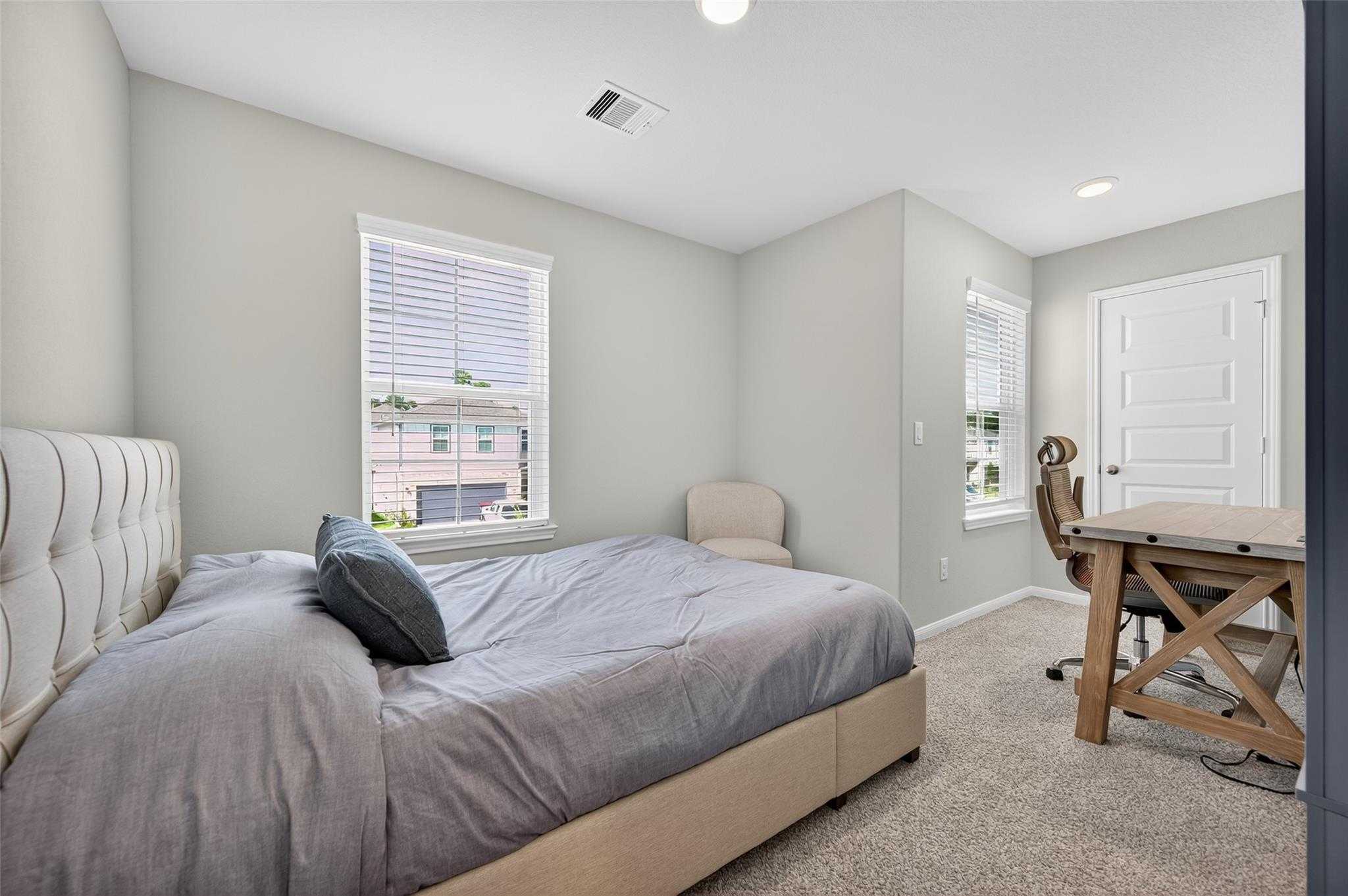 Inviting gray bedroom with tufted bed, wooden desk, armchair, and window views in Davidson Homes The Brazos E, Magnolia TX