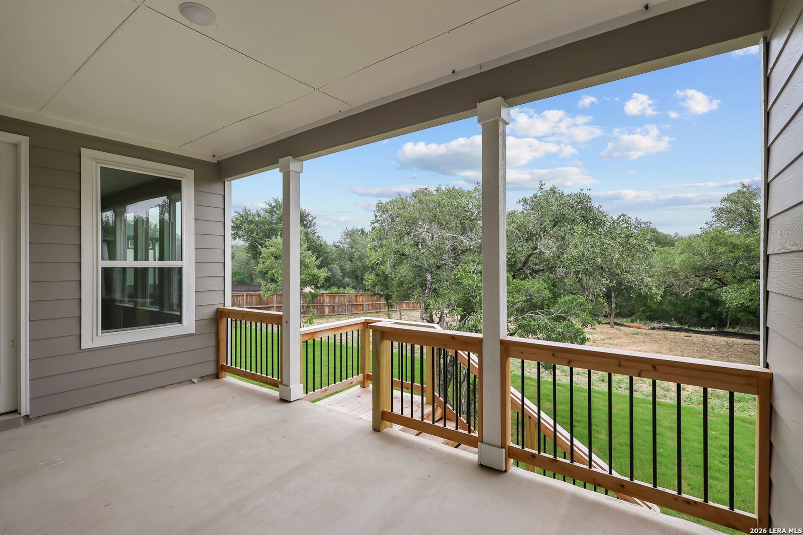 Covered porch with wooden deck, black iron railings, and stairs to green lawn amid oak trees in Davidson Homes The Belmont H, Castroville, Texas