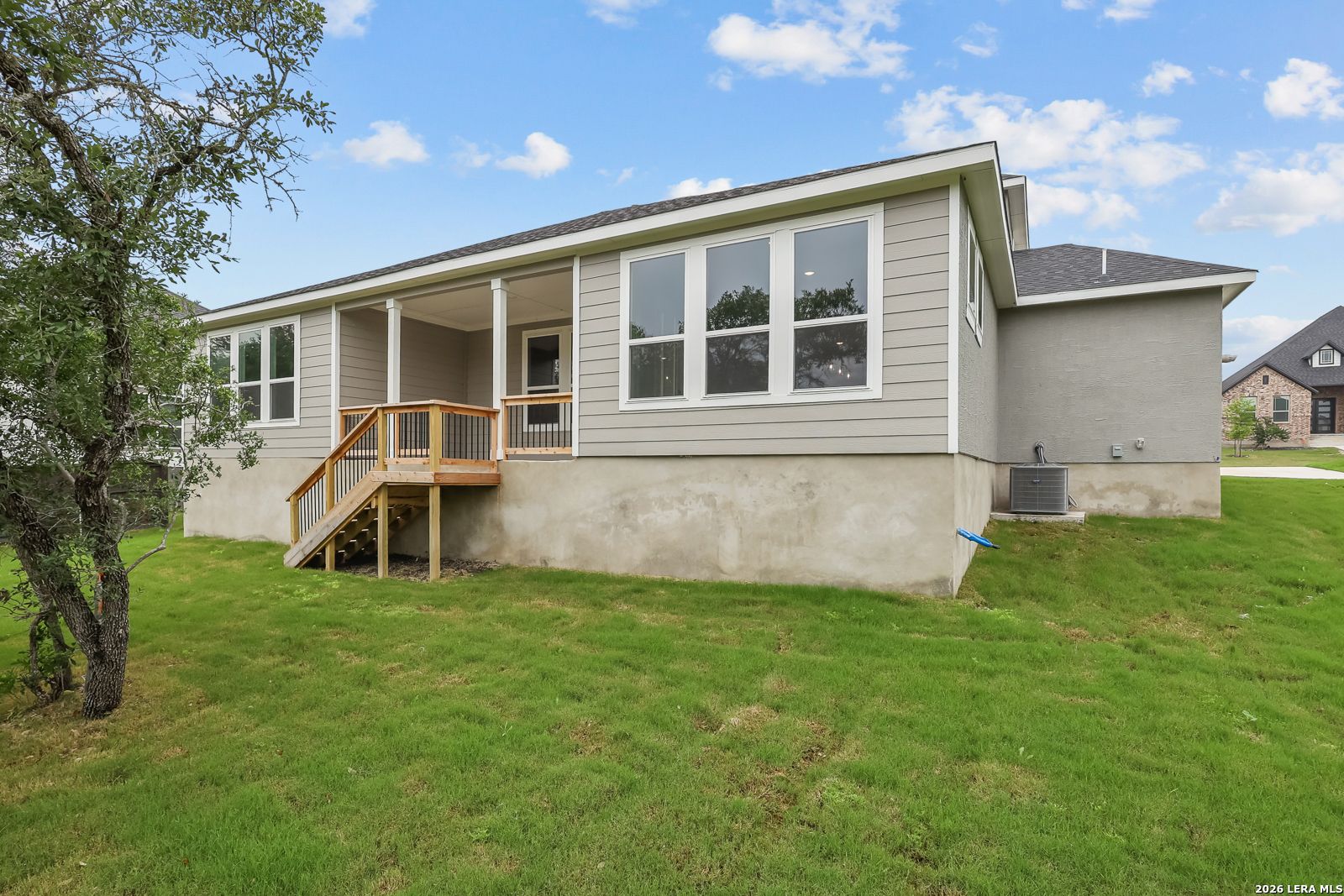Two-story Belmont H home side view with wooden deck stairs, large windows, and lush green yard in Potranco Oaks, Castroville, Texas