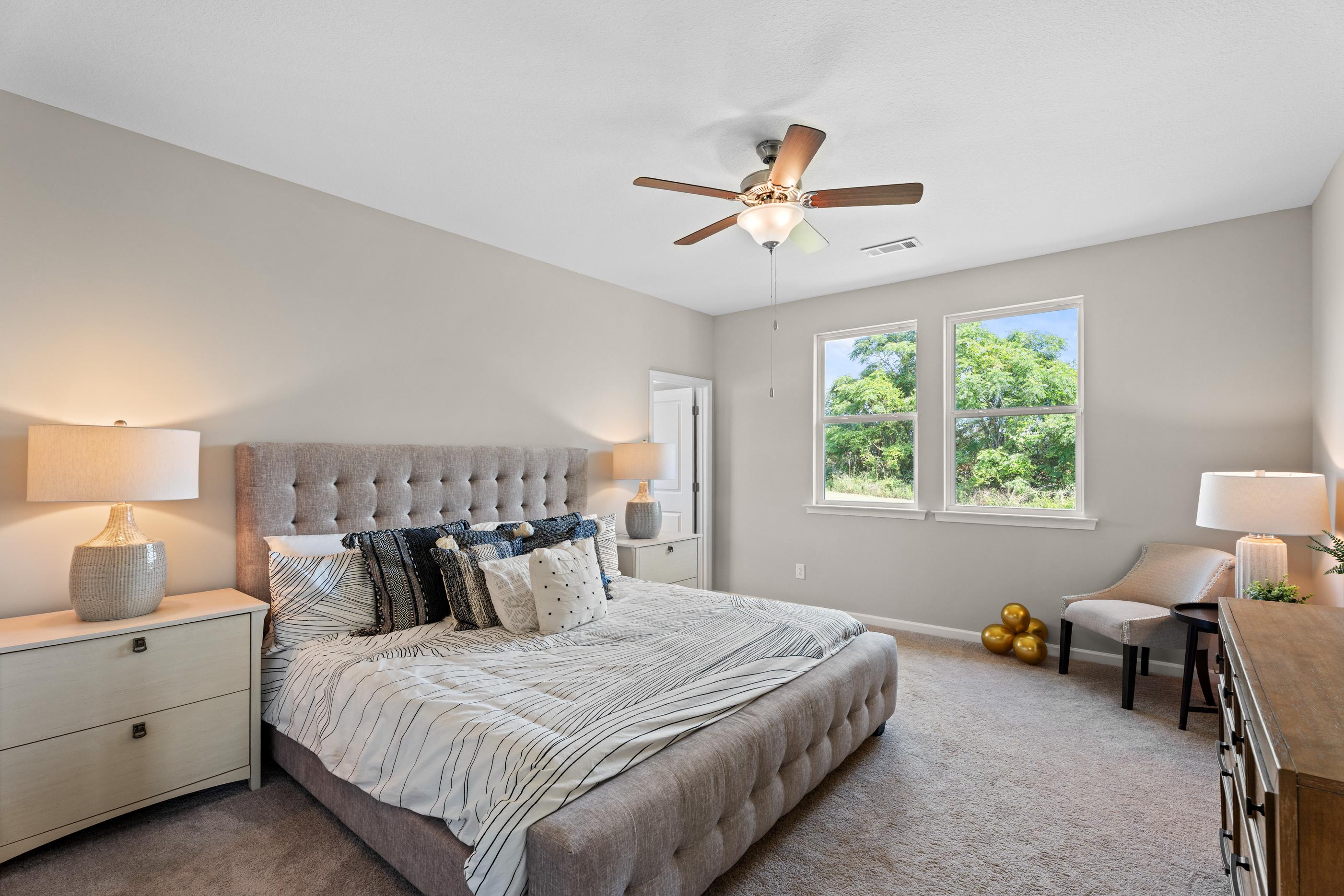 Spacious master bedroom in Ivy Glen Perry Georgia with tufted gray bed, ceiling fan, and large windows overlooking trees
