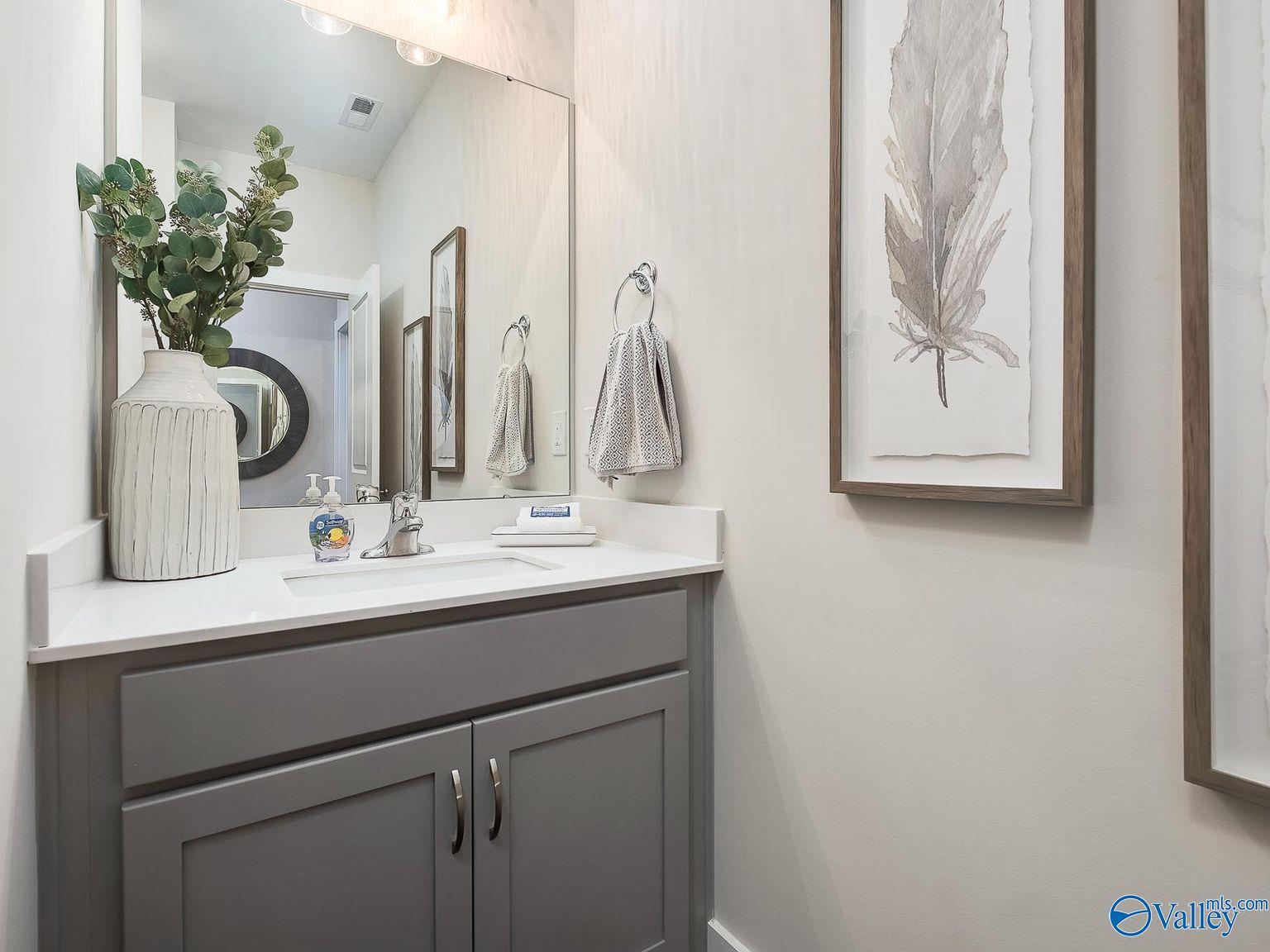 Modern powder room with gray vanity, white countertop sink, round mirror, eucalyptus vase, and feather art in The Everett by Davidson Homes, Toney, AL