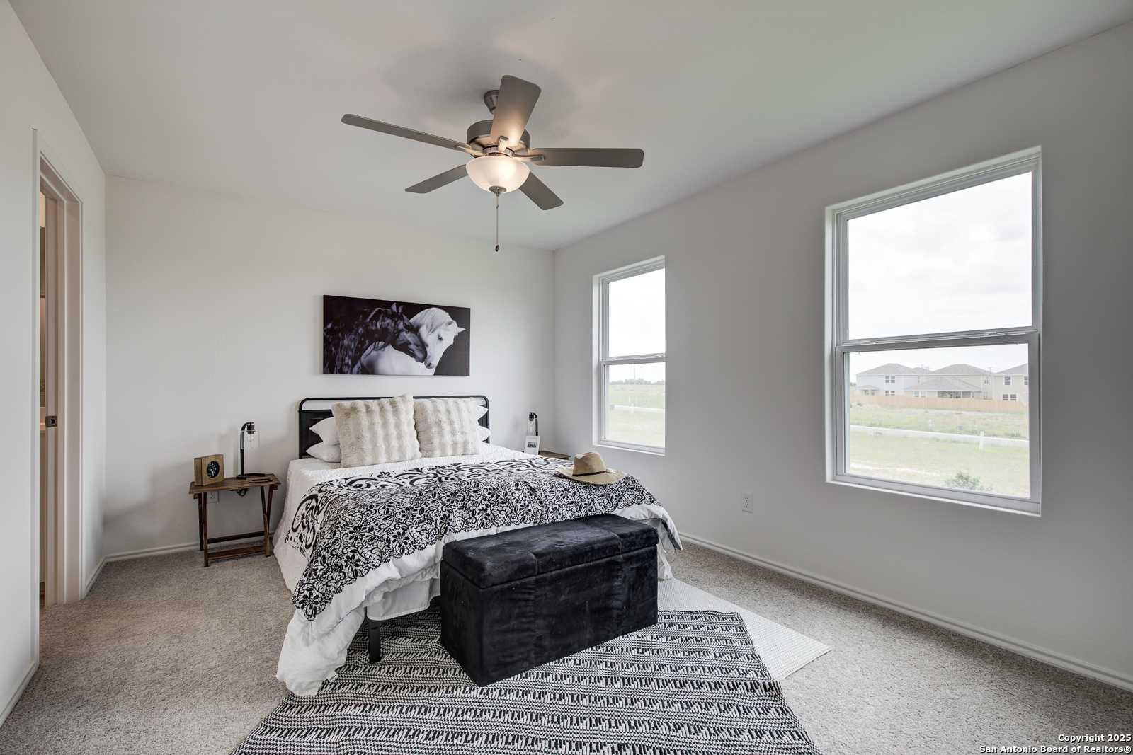 Spacious bedroom with king bed, patterned bedding, ceiling fan, large windows, and wall art in Davidson Homes The Trinity A, San Antonio