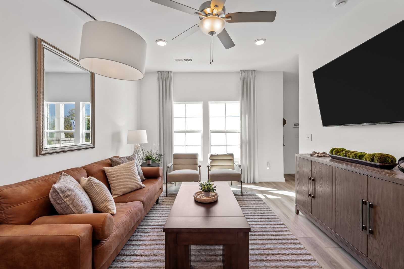 Cozy living room featuring brown leather sofa, mounted TV, ceiling fan, and large windows in Davidson Homes The Cumberland B, Gallatin, Tennessee