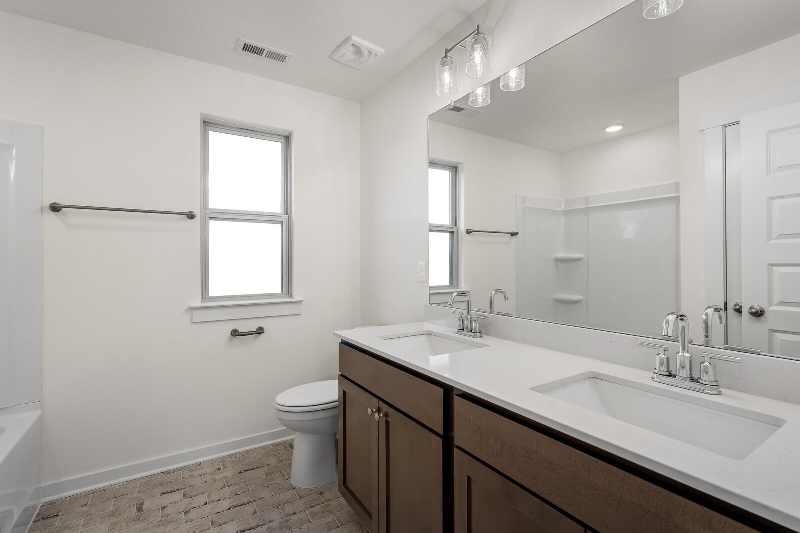 Modern bathroom with double vanity, white quartz counters, brown cabinets, glass shower, and brick tile floor in Davidson Homes Willow C, Gallatin, TN