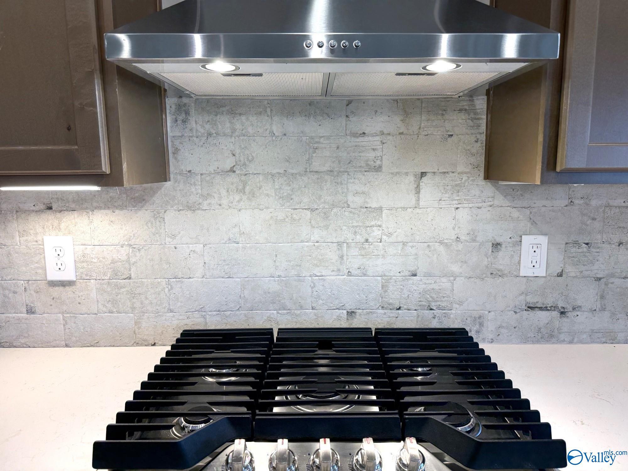 Stainless steel range hood and gas cooktop with textured tile backsplash in The Rockford floor plan, Hartselle, Alabama.