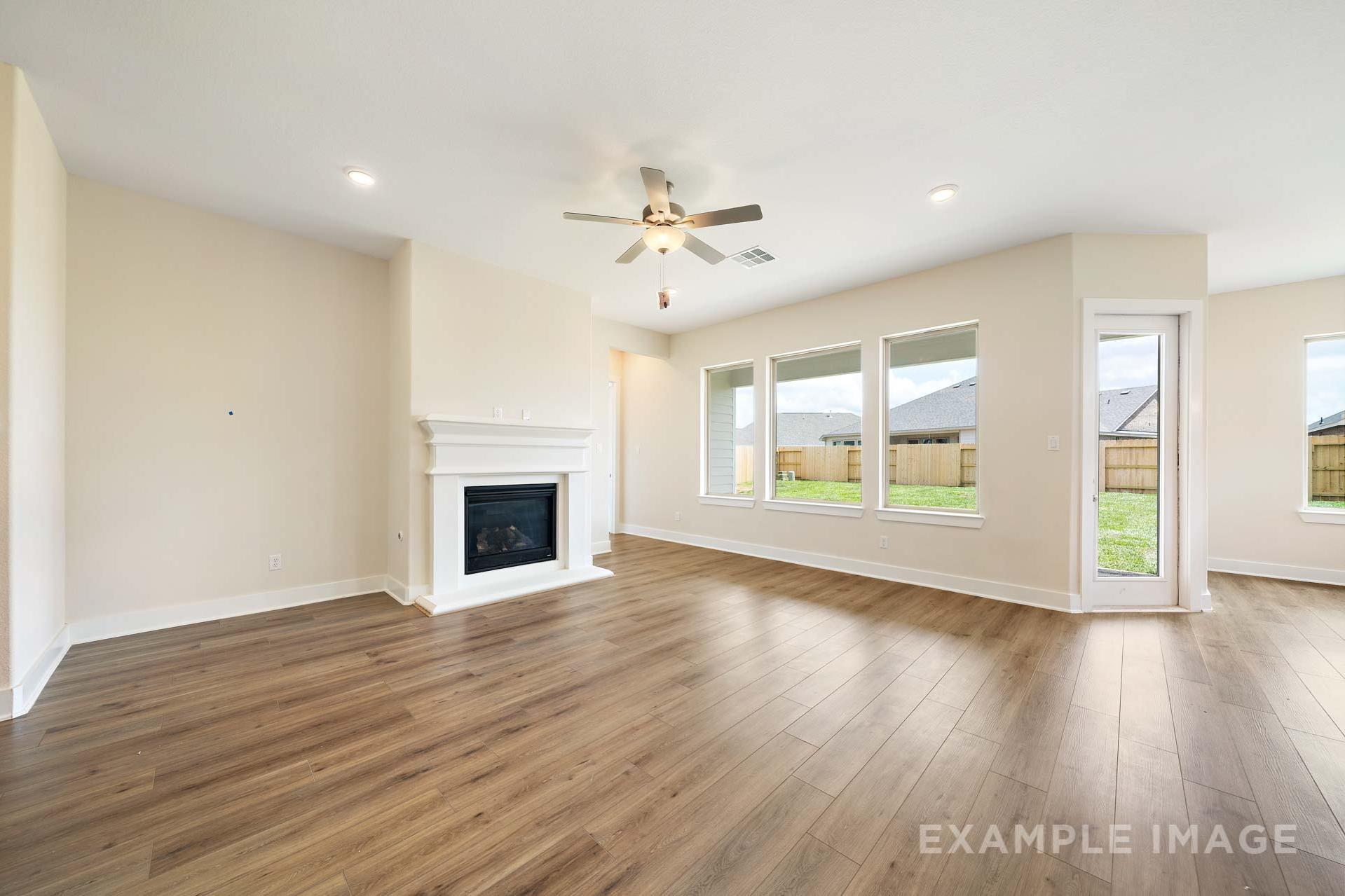 Spacious open-concept living room in The Diana C home with white fireplace, hardwood floors, and large windows overlooking backyard