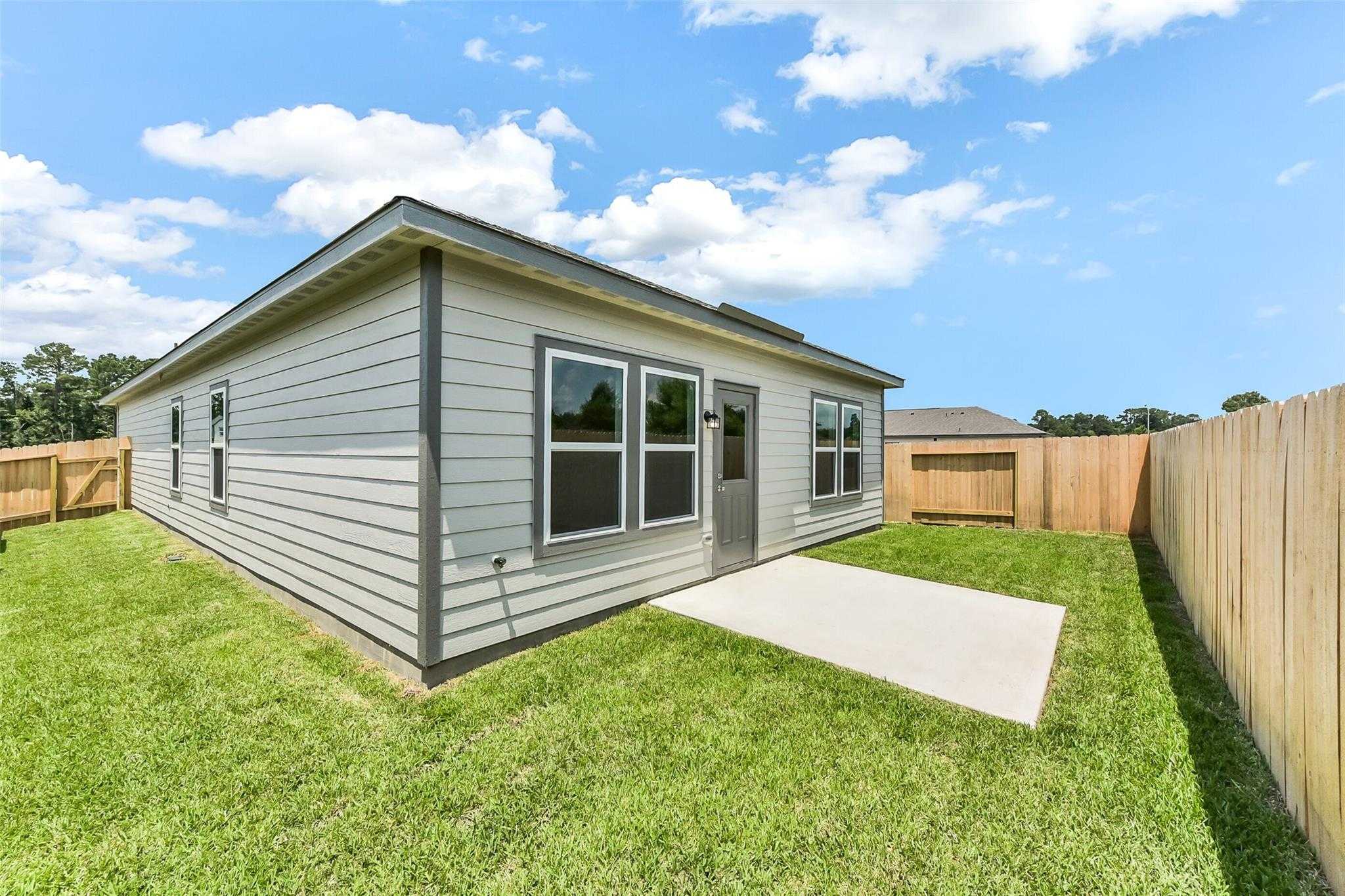 Gray single-story Davidson Homes The Frio F with concrete patio, lush green lawn, and wooden privacy fence in The Villages at WestPointe, Dayton, Texas