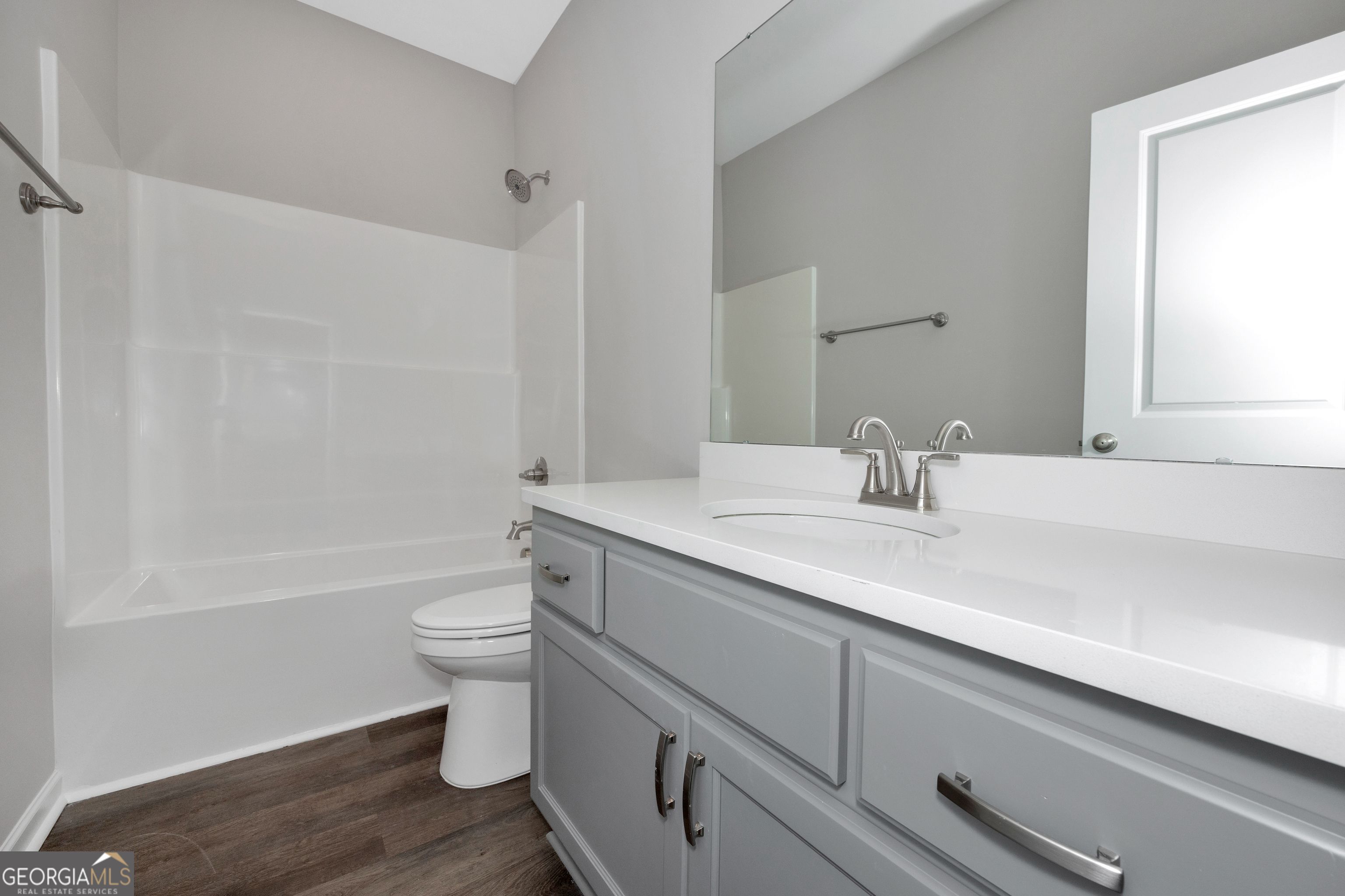 Modern guest bathroom with white tub-shower combo, gray vanity, large mirror, wood floors in Evermore Homes The Luna, Ivy Glen, Perry, Georgia