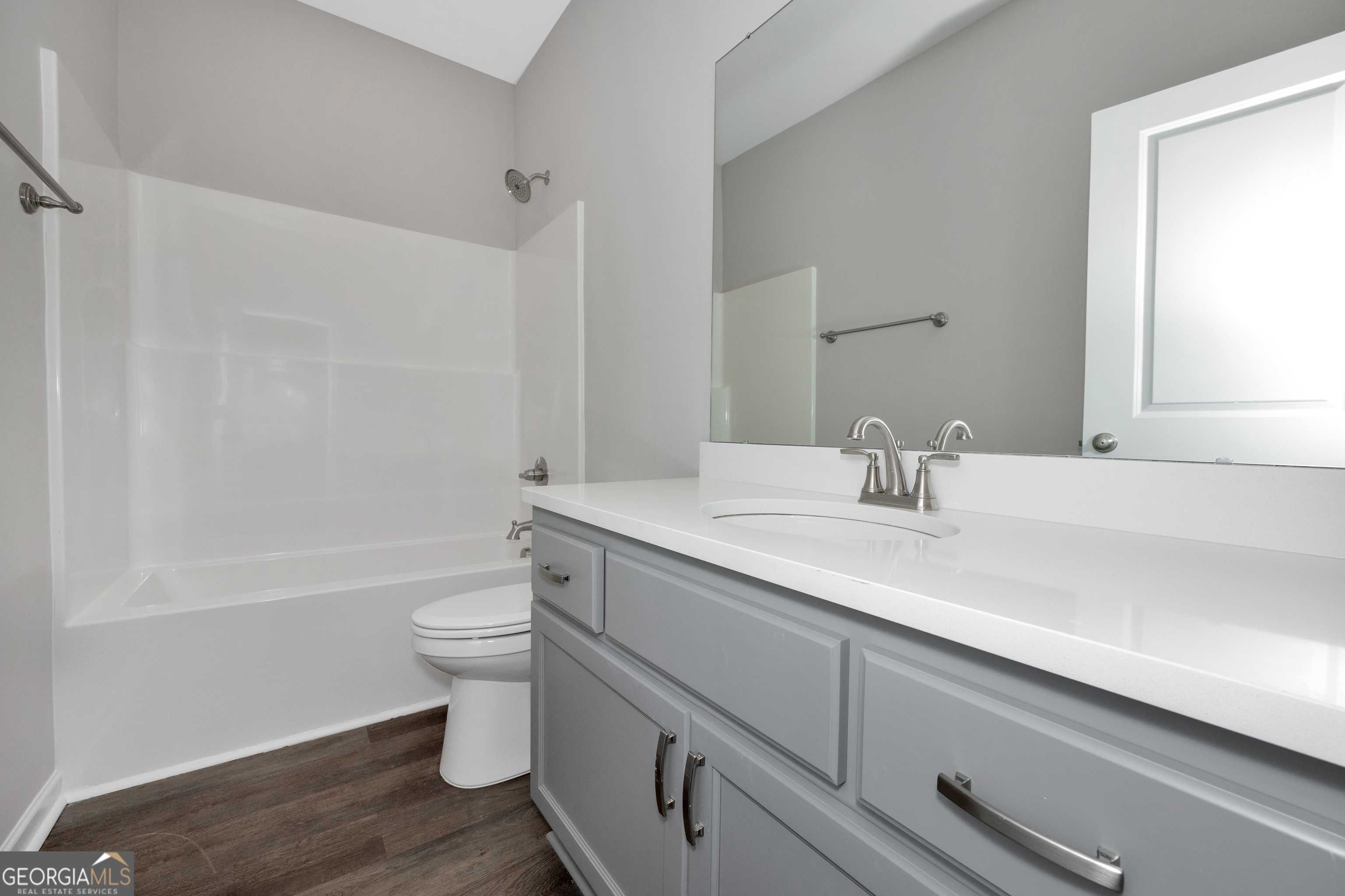 Modern guest bathroom with white tub-shower combo, gray vanity, large mirror, wood floors in Evermore Homes The Luna, Ivy Glen, Perry, Georgia