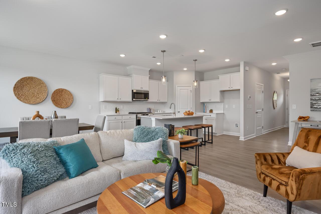 Open-concept living room with white sectional sofa, turquoise pillows, wooden coffee table, and adjacent kitchen in Davidson Homes The Warren, Fuquay-Varina, NC