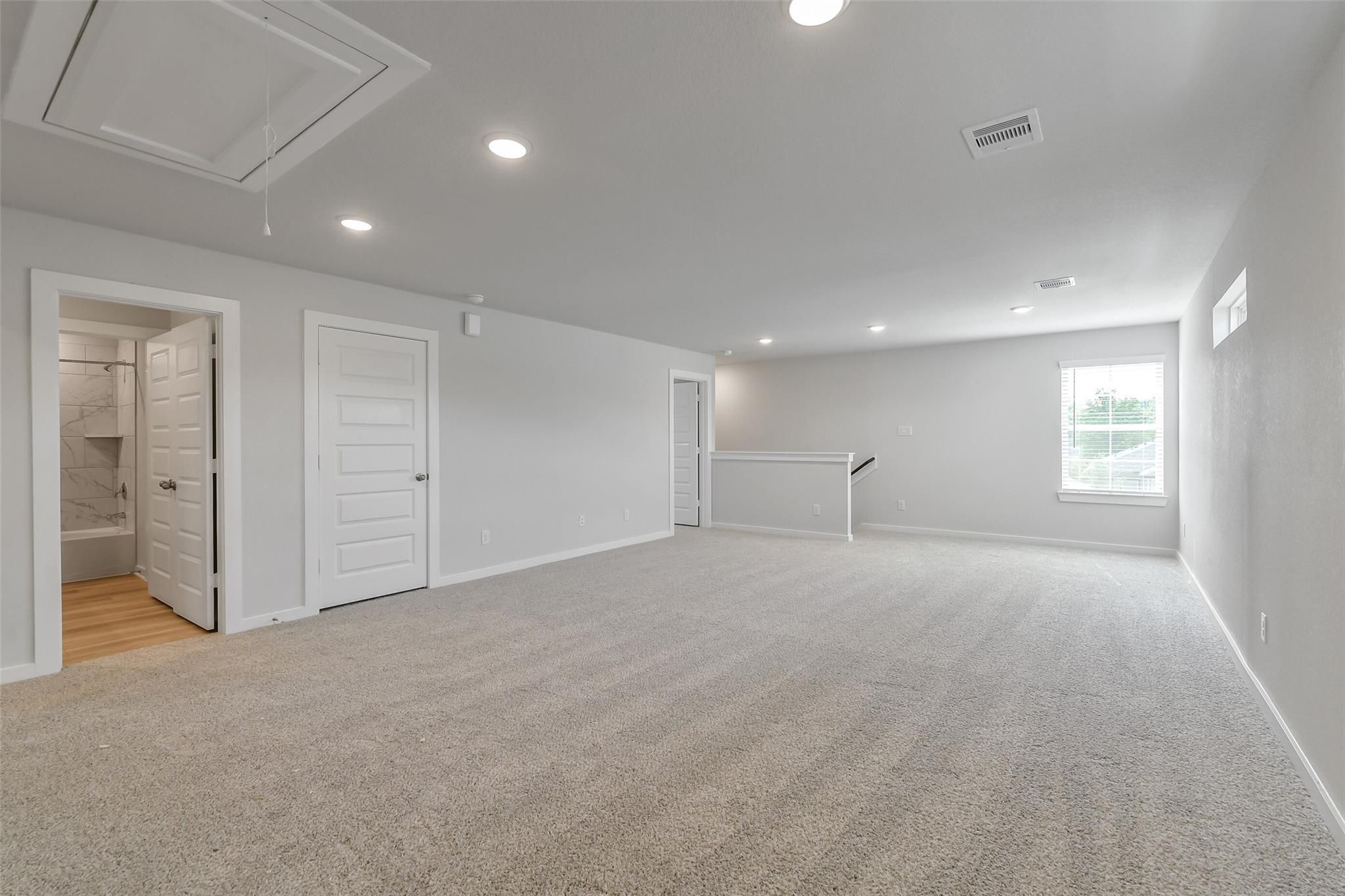 Spacious upstairs bonus room with beige carpet, adjacent bathroom tub, window, and railing in 5-bedroom San Marcos E home, Conroe, Texas
