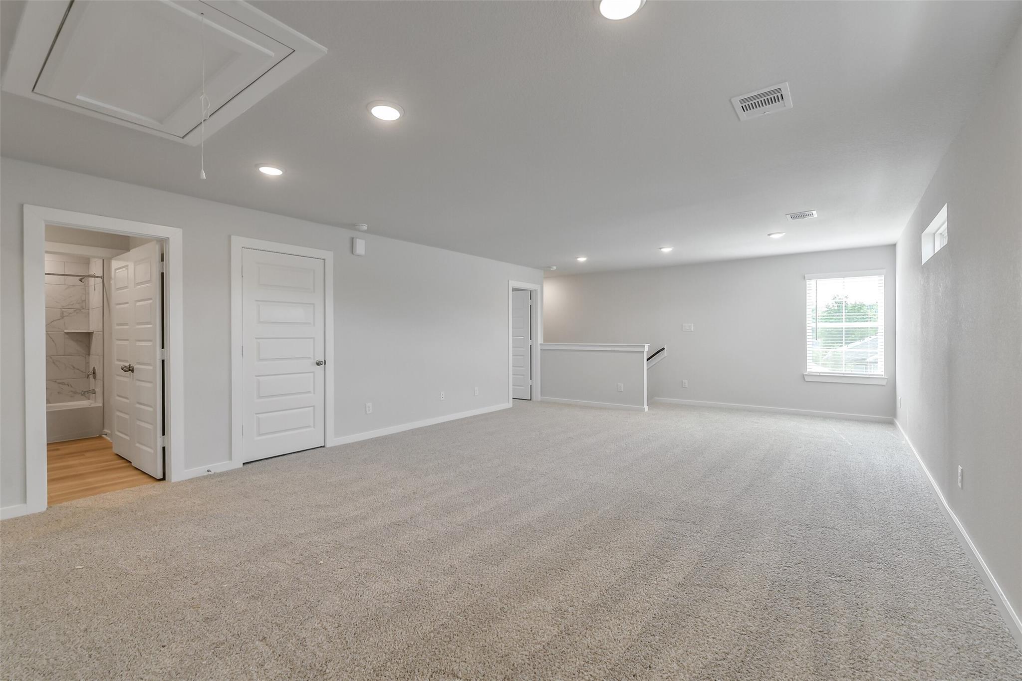 Spacious upstairs bonus room with beige carpet, adjacent bathroom tub, window, and railing in 5-bedroom San Marcos E home, Conroe, Texas