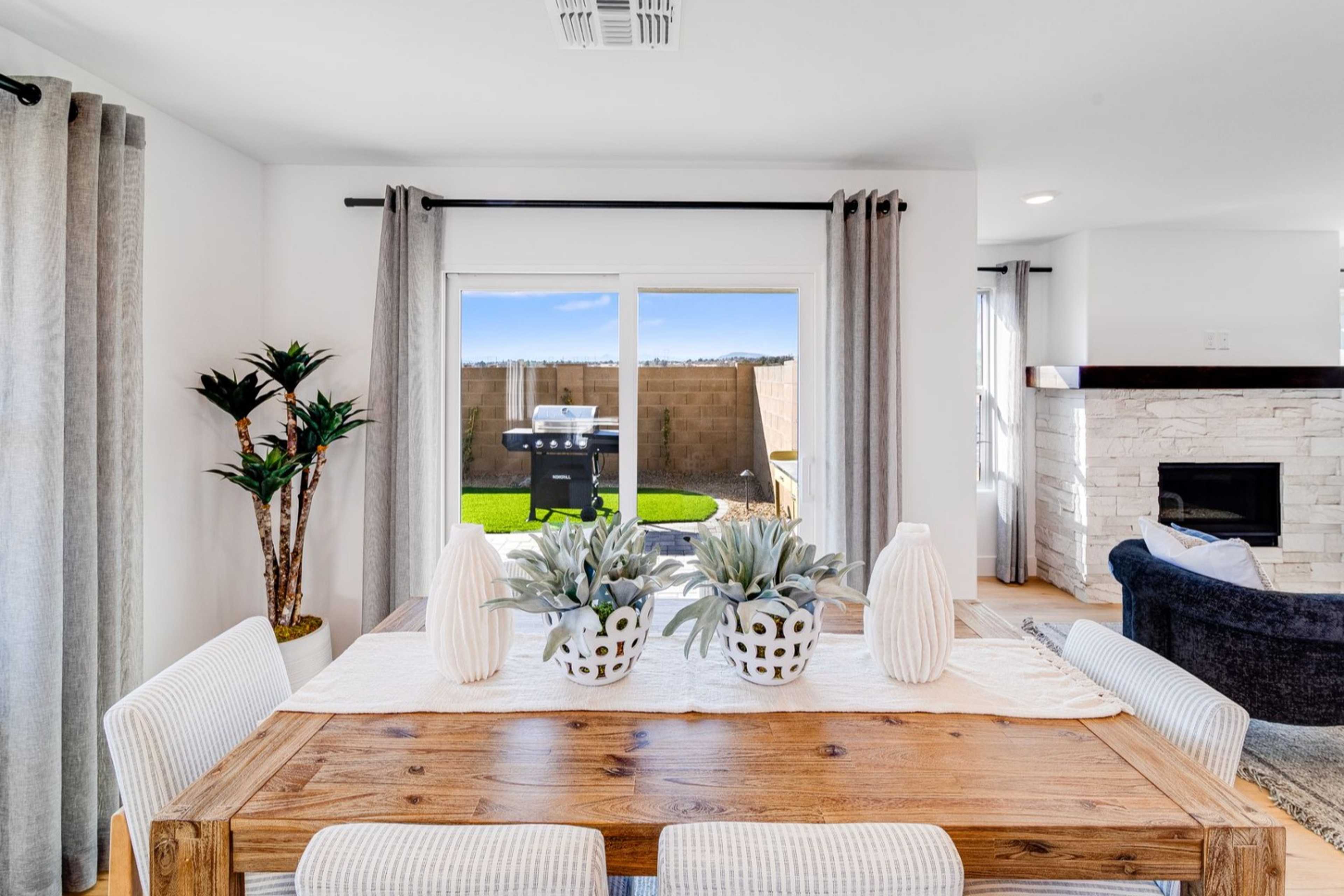 Spacious dining area in The Wilmington B with wooden table, white chairs, potted plants, large windows to backyard patio, stone fireplace, curved sofa