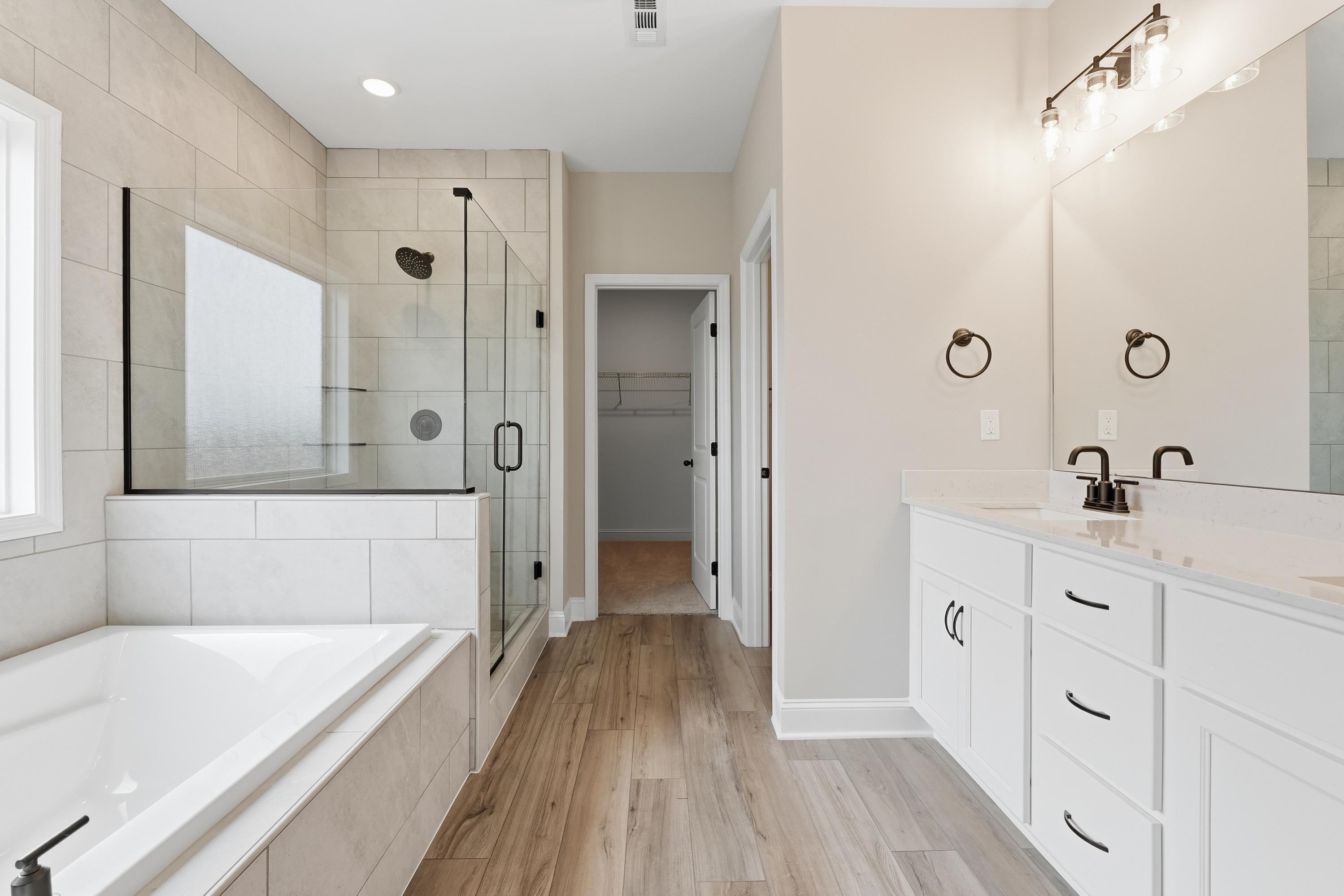 Spacious master bathroom in The Rockford with frameless glass shower, freestanding tub, double white vanity, and hardwood floors