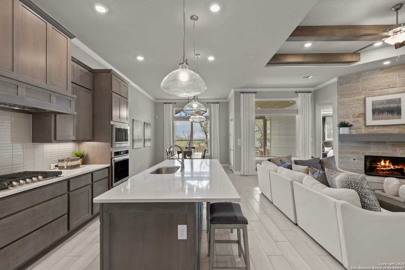 Open-concept kitchen with white island, stainless appliances, and living room fireplace overlooking patio in Davidson Homes The Garner A, Castroville, Texas