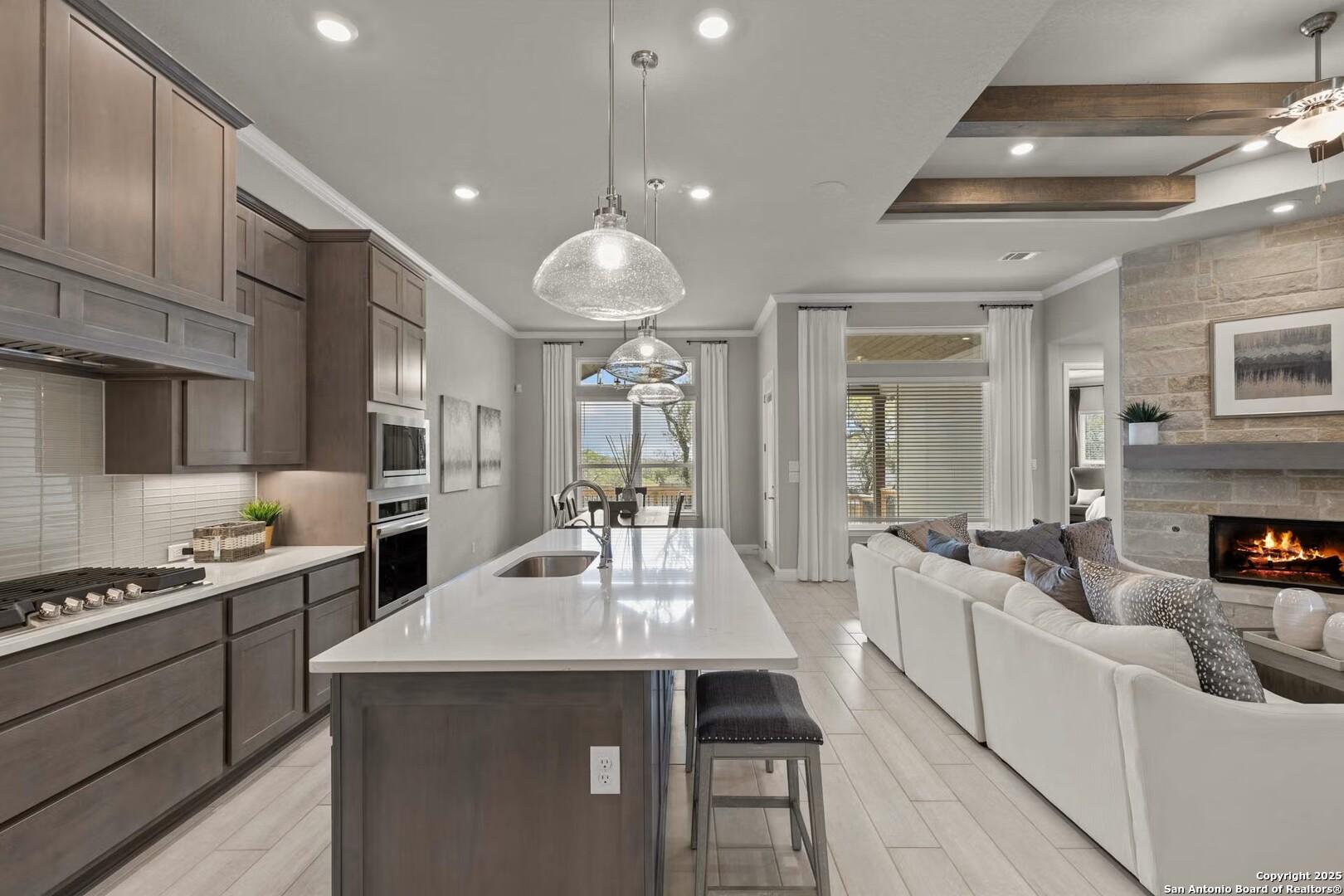 Open-concept kitchen with white island, stainless appliances, and adjacent living room featuring fireplace and backyard view in The Garner B, Castroville Texas