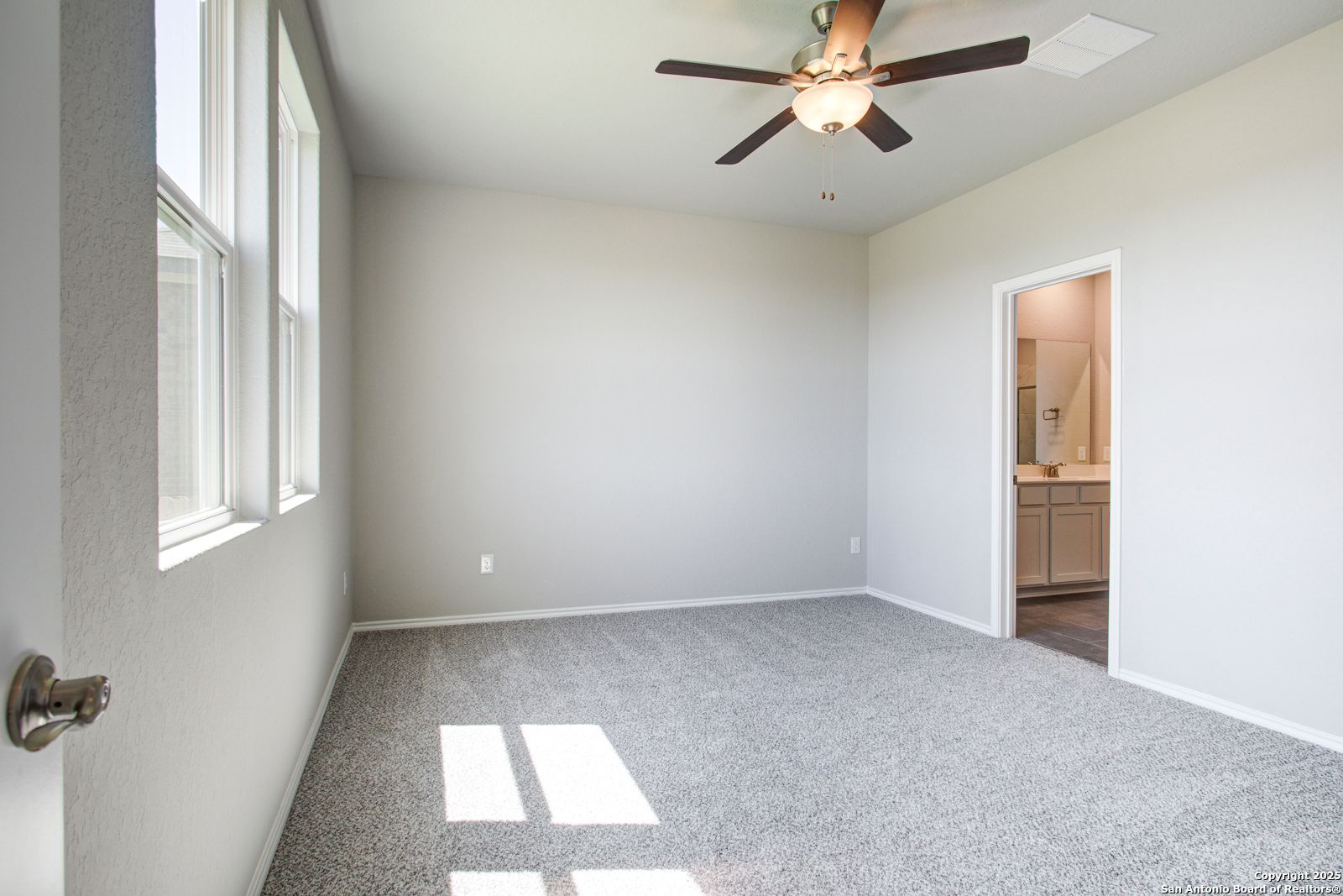 Bright bedroom with ceiling fan, gray carpet, large windows, and en-suite bathroom in Davidson Homes The Douglas G, San Antonio