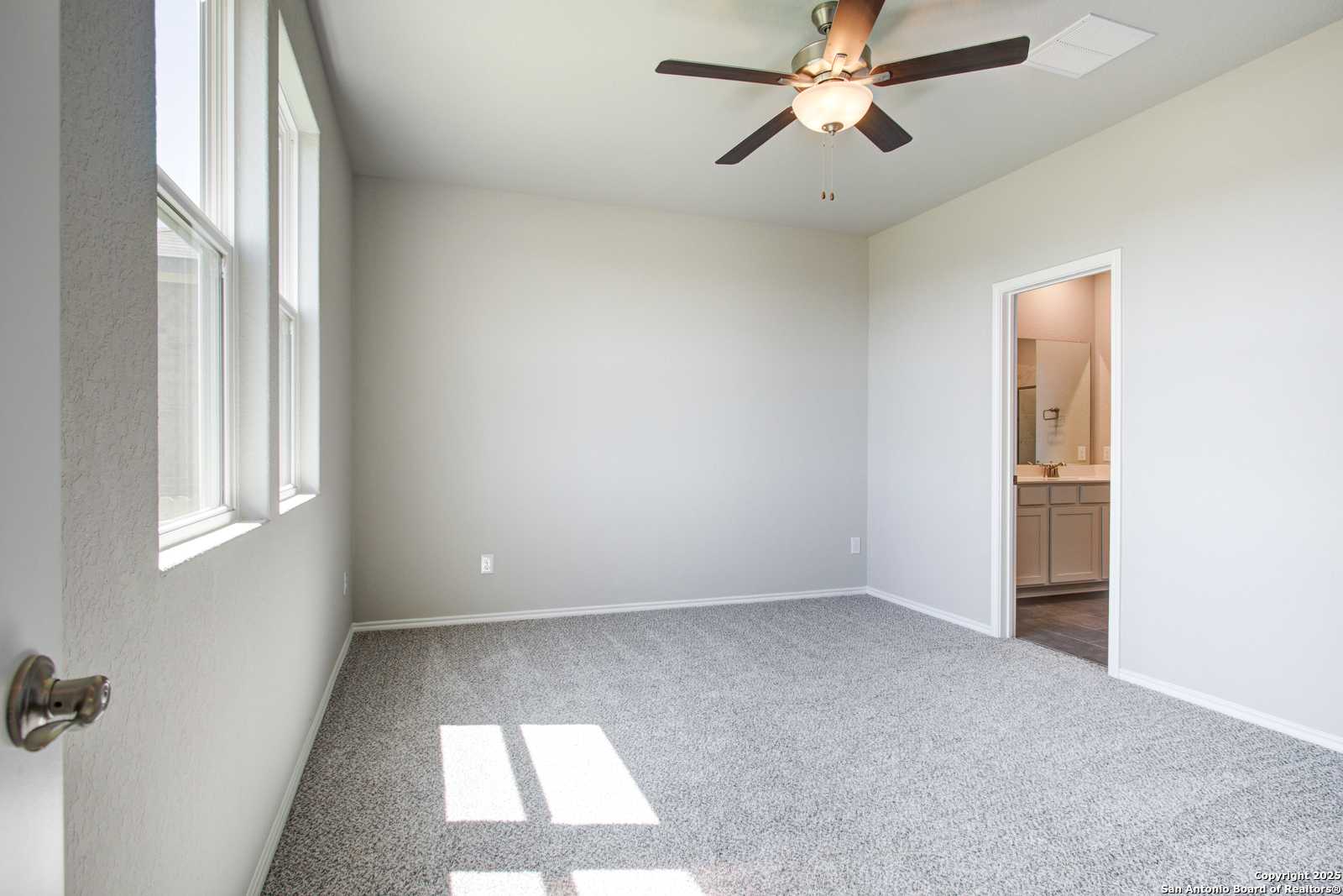 Bright bedroom with ceiling fan, gray carpet, large windows, and en-suite bathroom in Davidson Homes The Douglas G, San Antonio